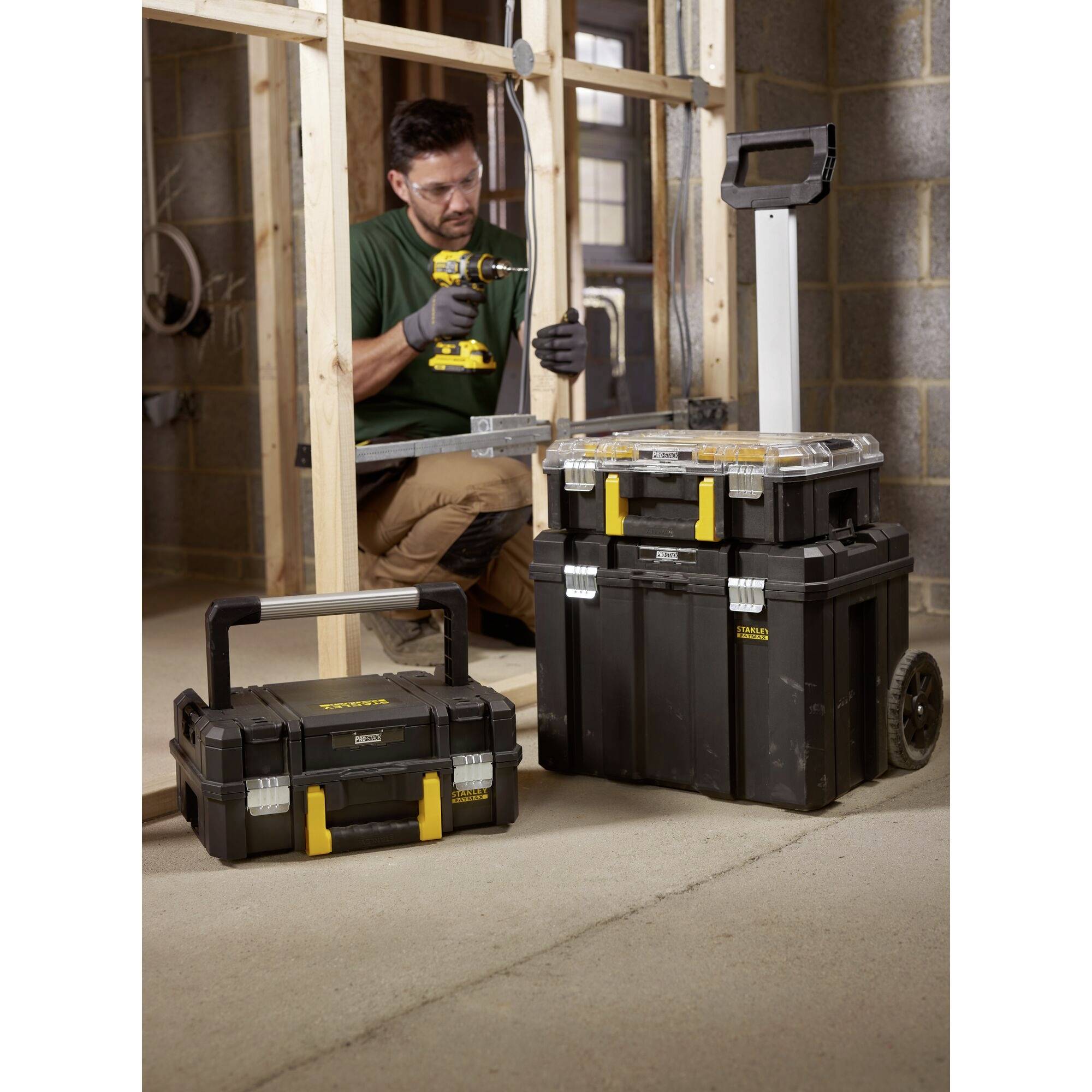 A worker in an unfinished room is using a drill. In the foreground, two black toolboxes with handles and yellow locks are positioned.