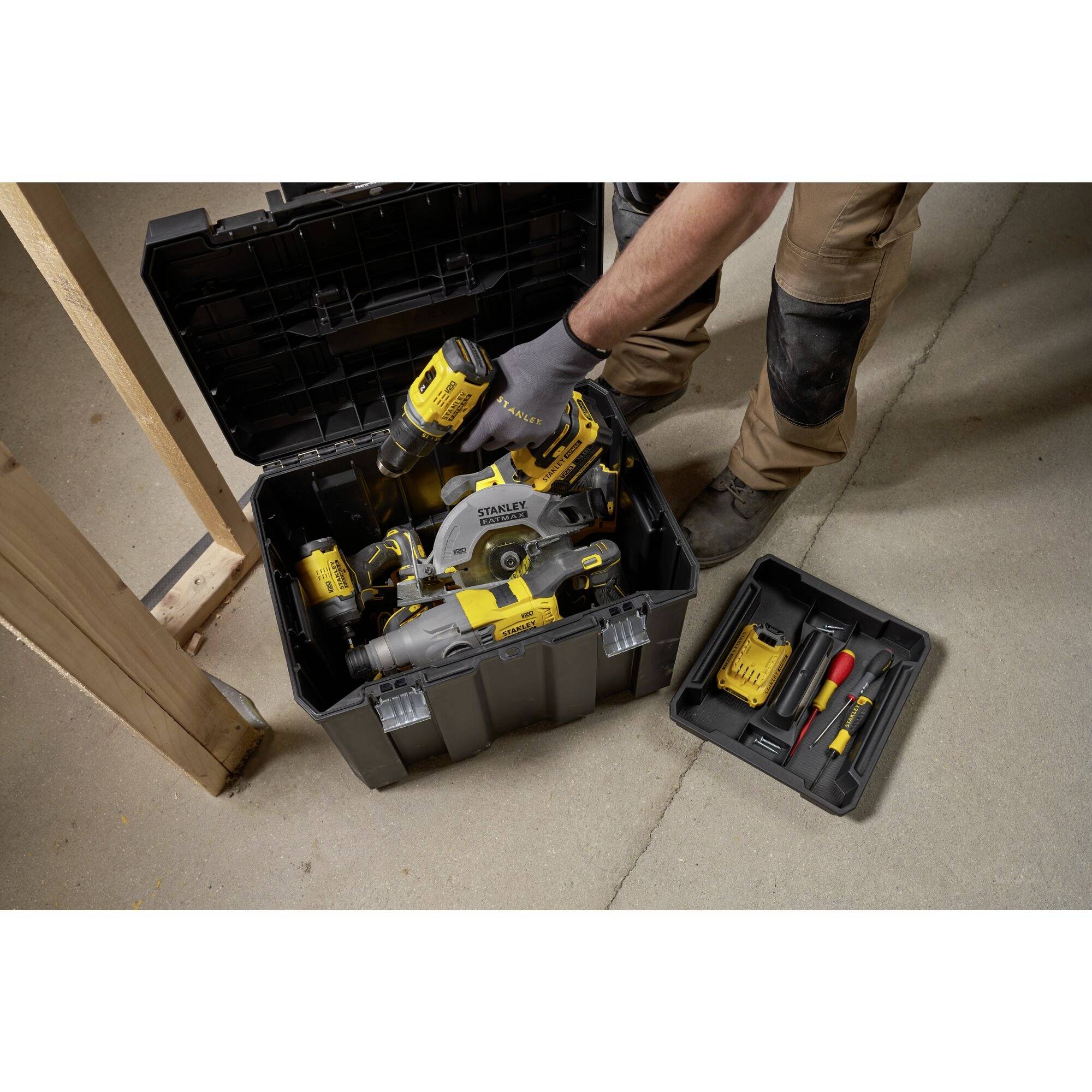 A person is leaning over an open toolbox containing a yellow saw and accessories. A separate box with additional tools lies beside it.