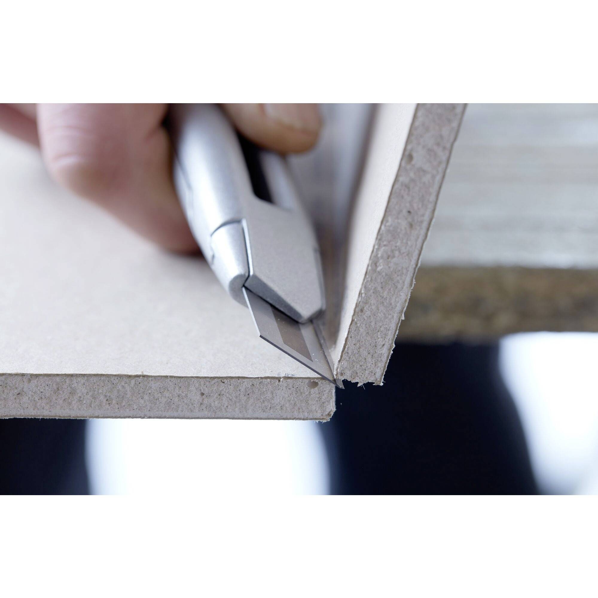 A hand cuts a straight line into a cardboard sheet with a craft knife on a table.