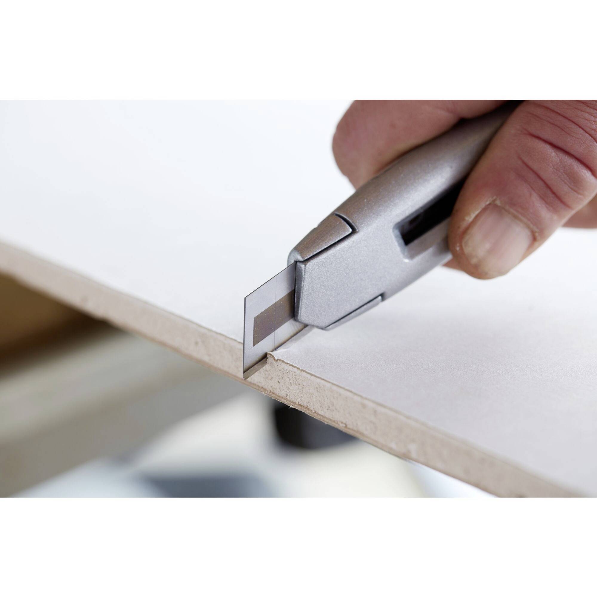 A hand cuts precise lines on a white cardboard using a silver utility knife along the edge of a table.