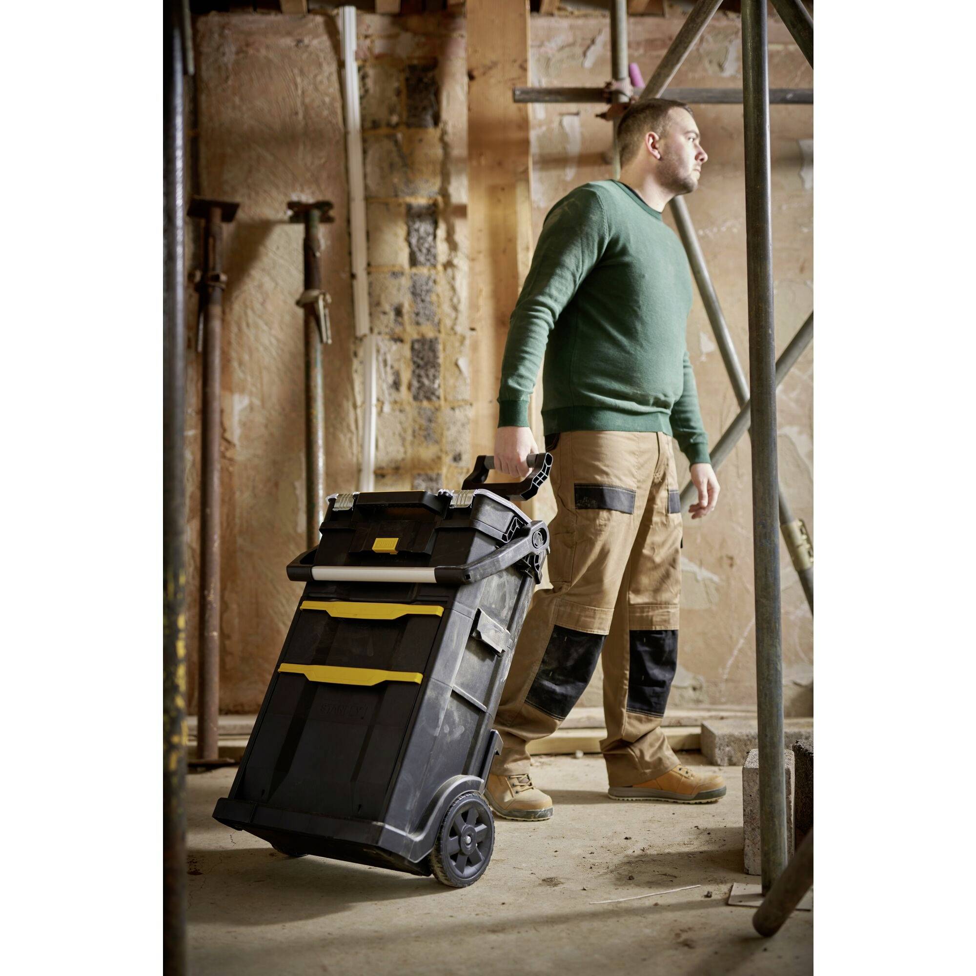 A man in workwear is pulling a large, black toolbox on wheels in an unfinished room.