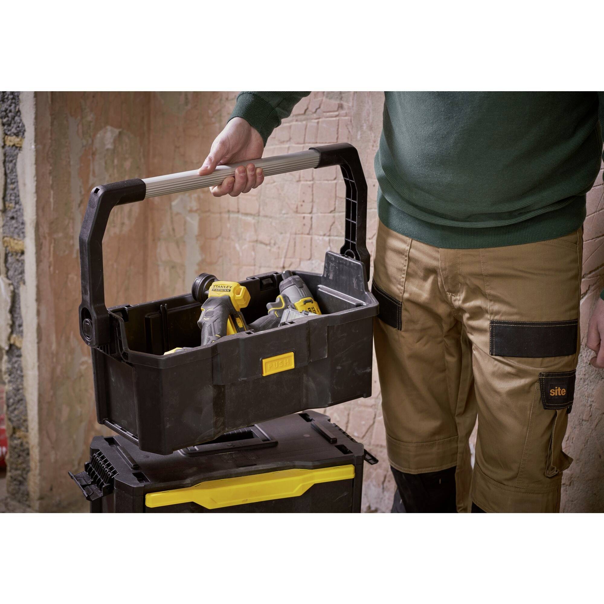A person wearing work trousers is holding a toolbox with several power tools, against a rustic, unfinished background.