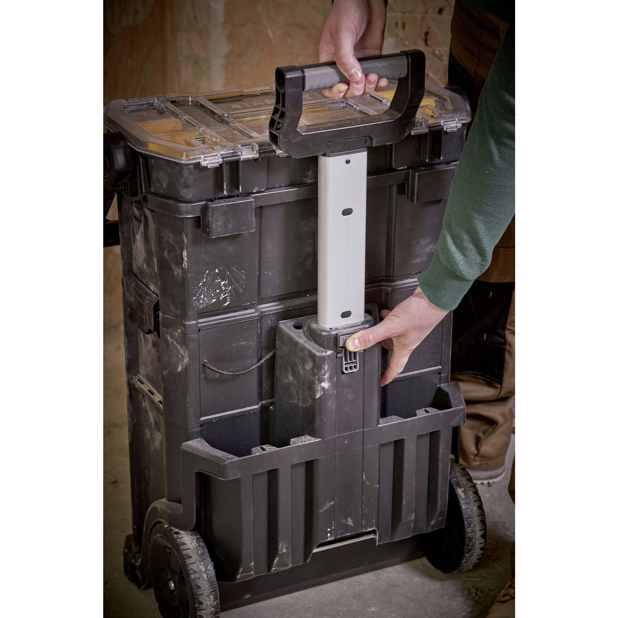 A person is holding a black tool trolley with a handle. The trolley has wheels and multiple compartments. The environment appears to resemble a construction site.