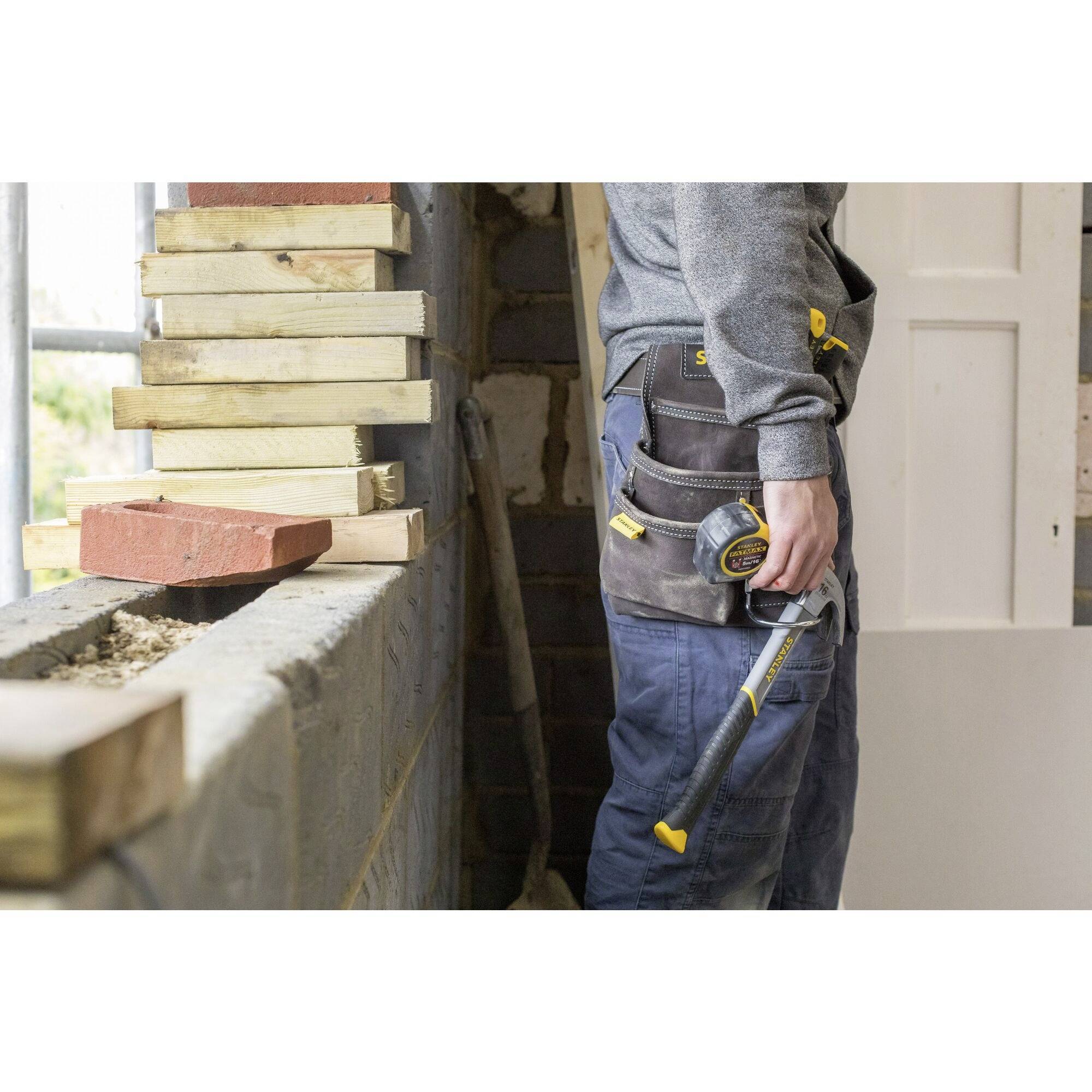A person is working on a construction site with stacks of wood and bricks. They are wearing site-appropriate clothing and have a tape measure and tools with them.
