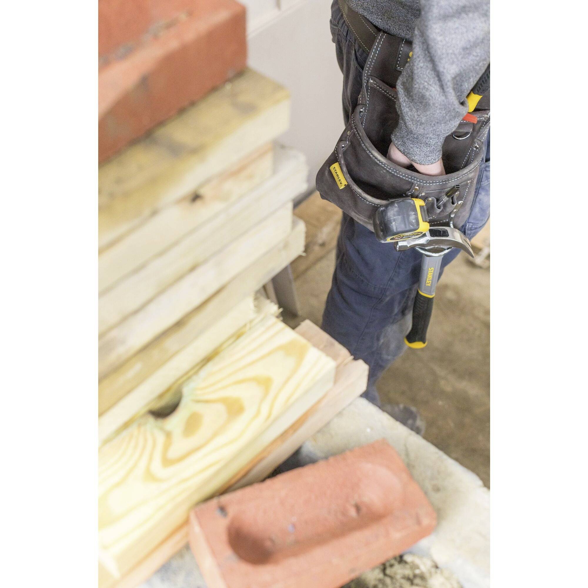 A person is standing next to a woodpile and brickwork. The individual is wearing a tool belt with a hammer, ready for construction work.