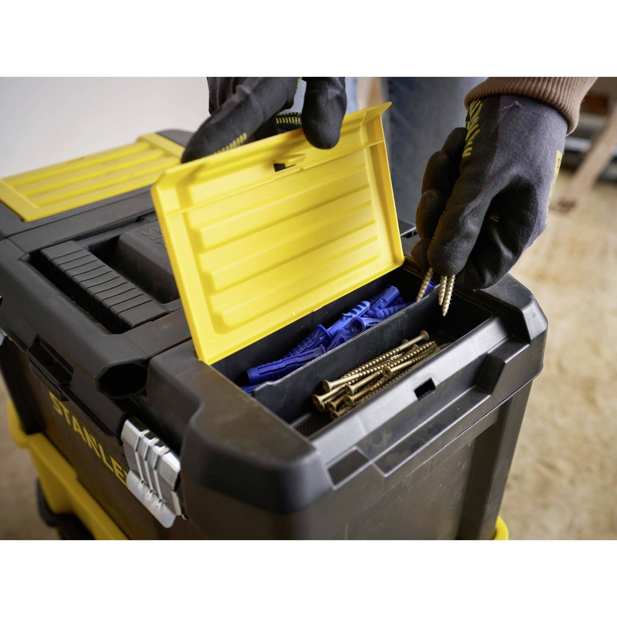 A person wearing gloves is placing screws into the upper compartment of a large black toolbox with a yellow lid.