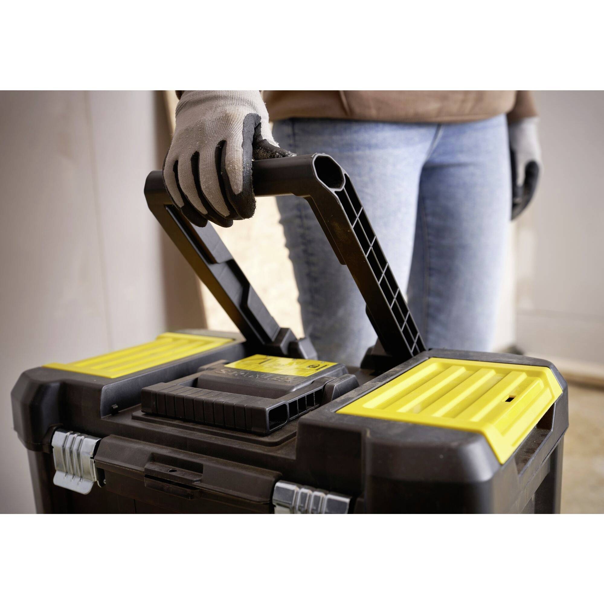 A person is holding a toolbox with a yellow lid handle. They are wearing gloves and jeans. The scene is located in an indoor setting.