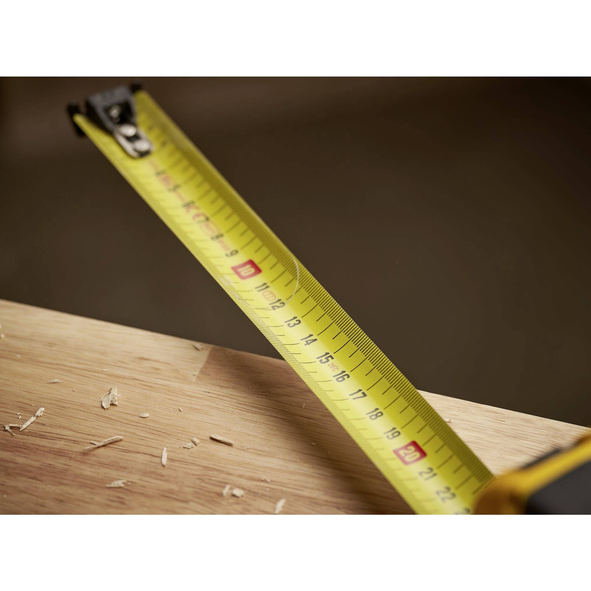 A yellow tape measure with centimetre markings is held over a wooden board scattered with wood shavings.