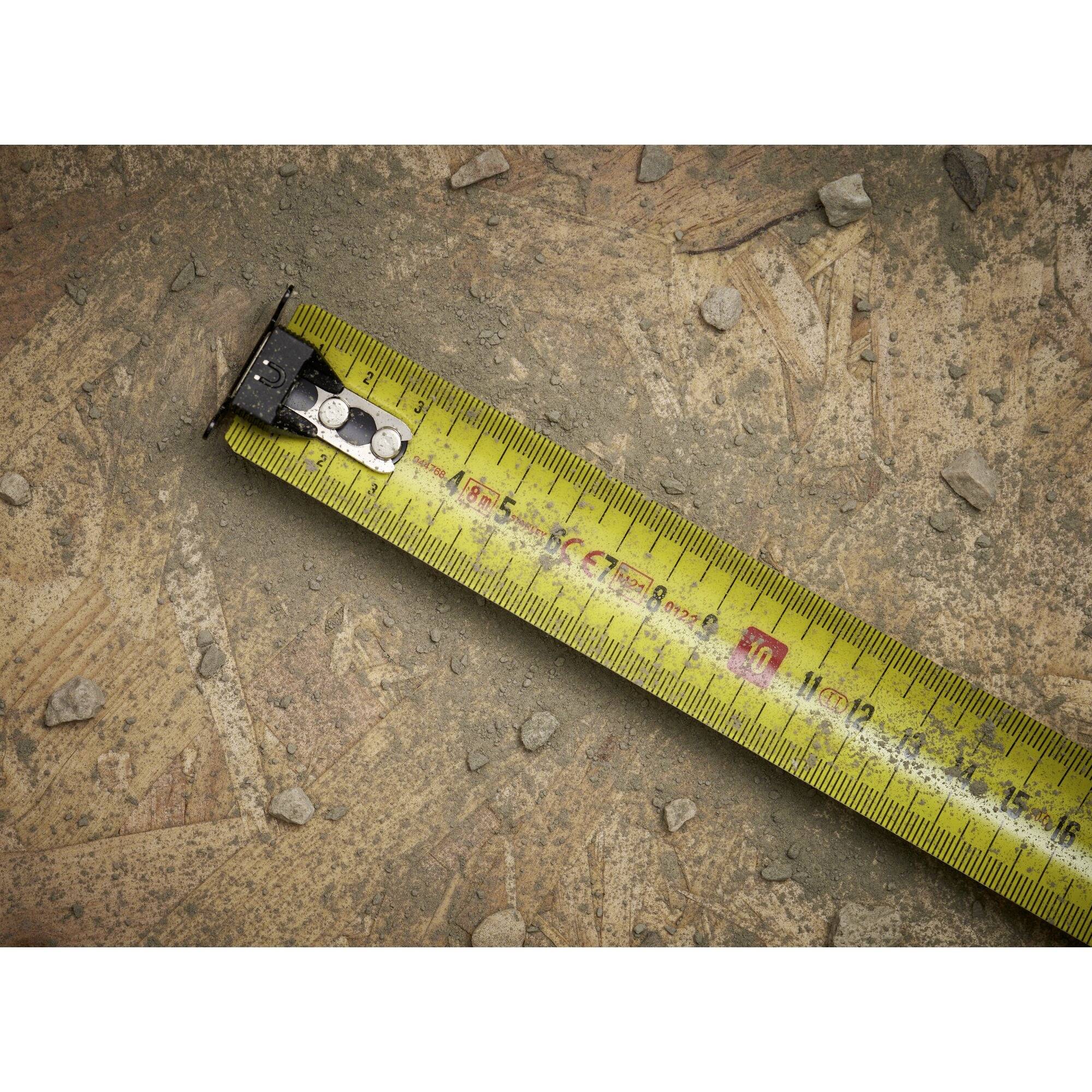 A yellow tape measure lies on a dusty floor with small stones. It is partially unrolled and indicates how something is being measured.