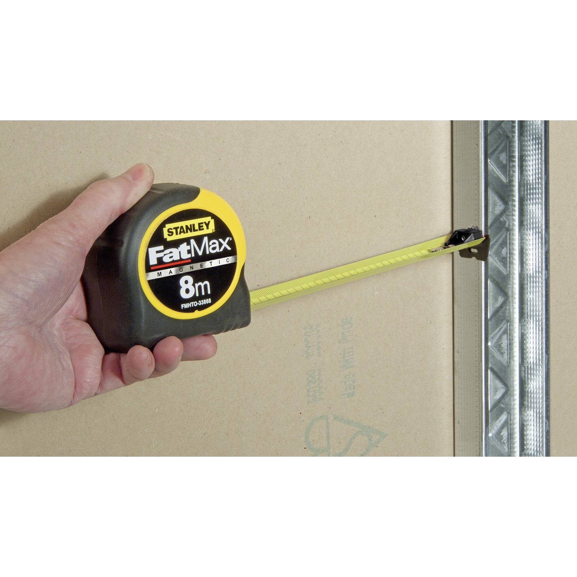 'A hand is holding an 8-metre Stanley FatMax tape measure, which is attached to a metal edge to measure a wall length.'