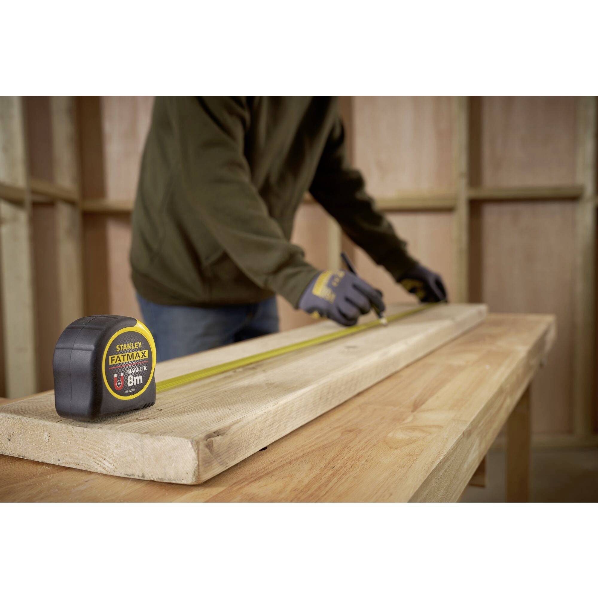A person is measuring a wooden board with a tape measure on a workbench, surrounded by unfinished wooden walls.