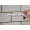 A hand is holding a razor blade with the inscription 'Carbide'. The background shows a tiled wall.