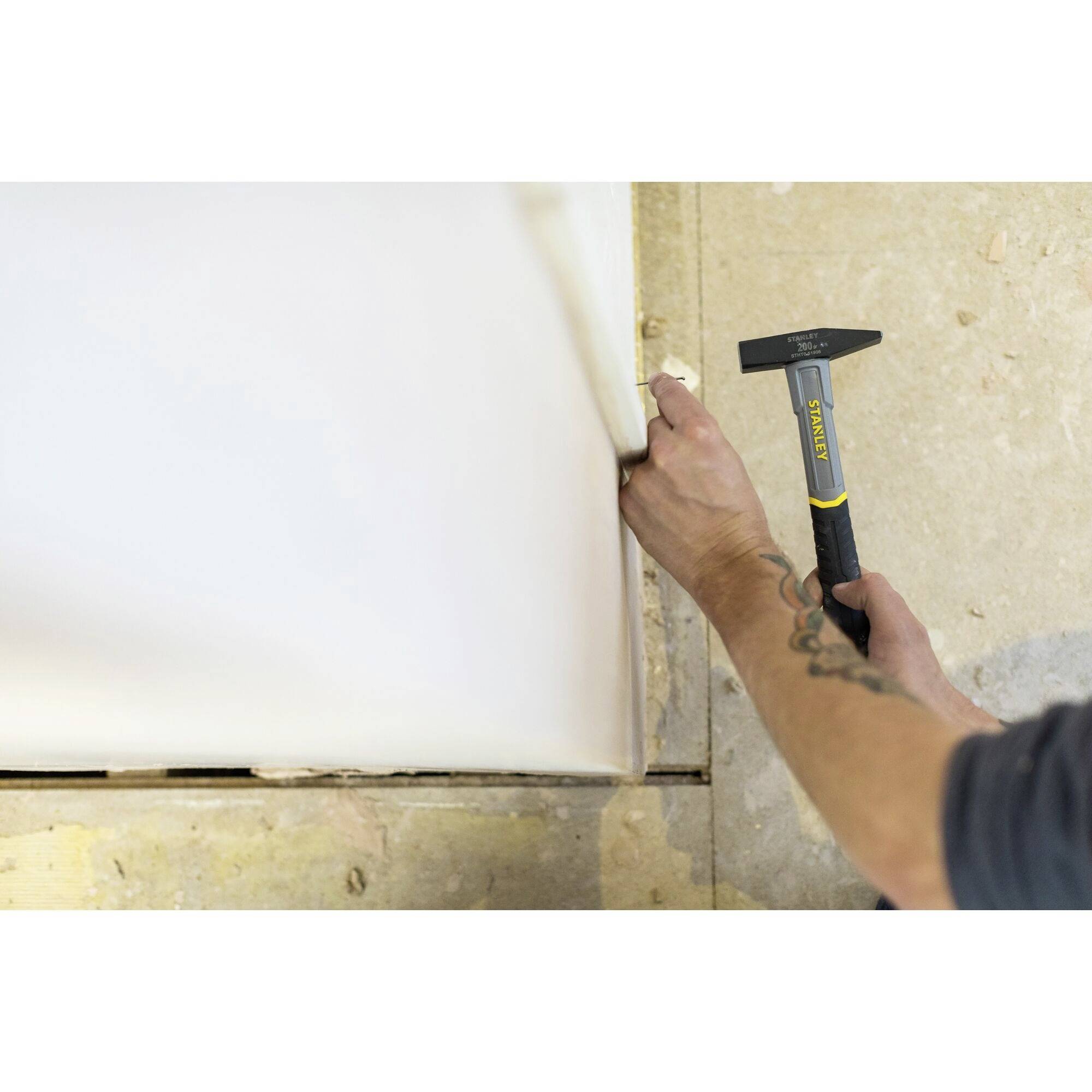 A person is using a hammer to work on a wall. The surroundings show a construction site floor and a light wall colour.
