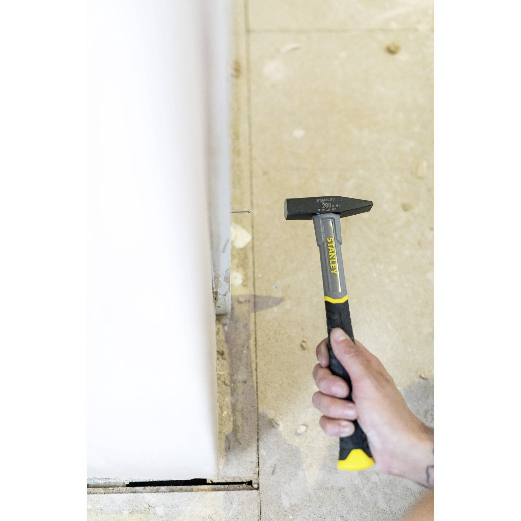 A person is holding a hammer and working at the bottom edge of a wall in a room with a concrete floor.