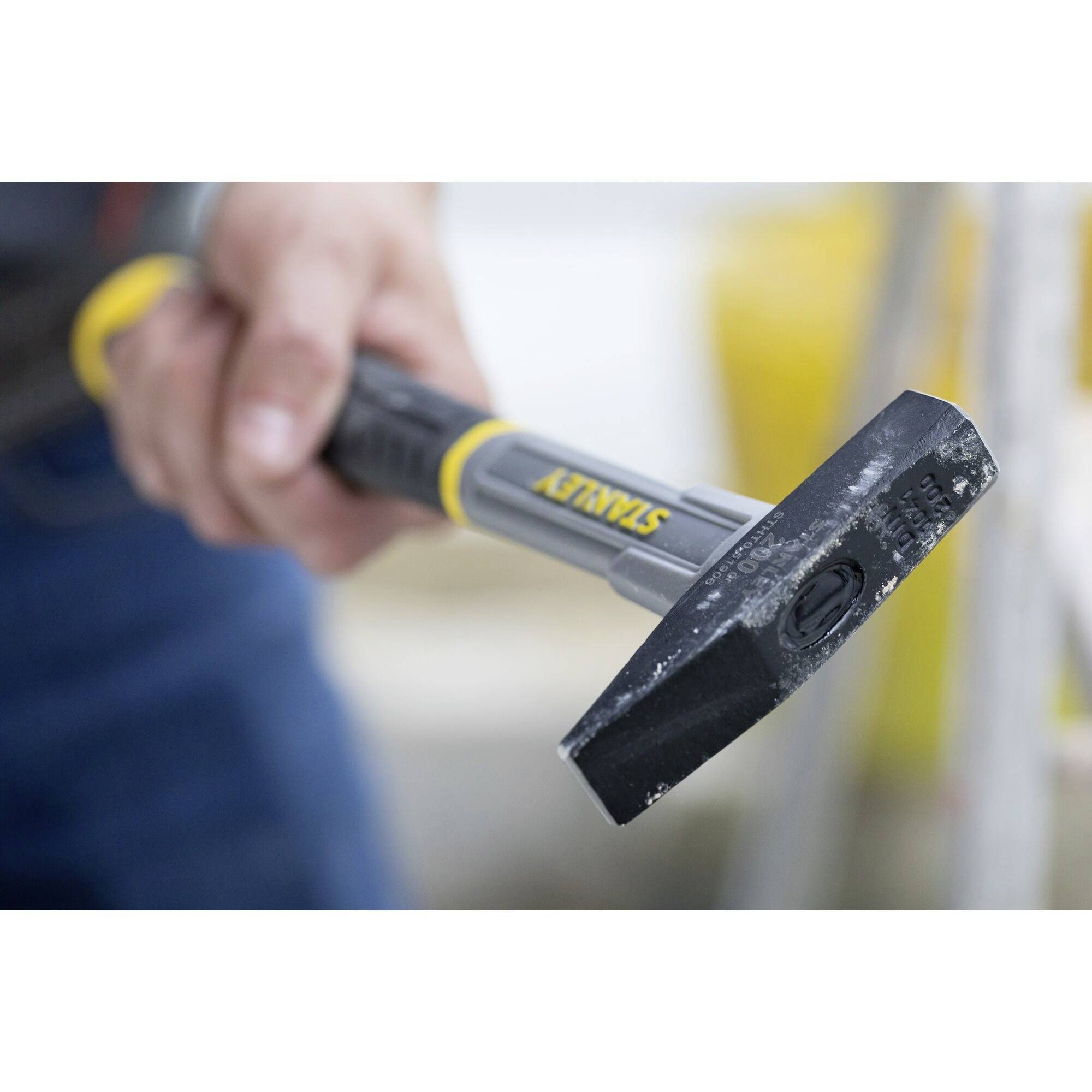 A hand is holding a black hammer with a yellow handle. The focus is on the hammer head. Background is blurred.