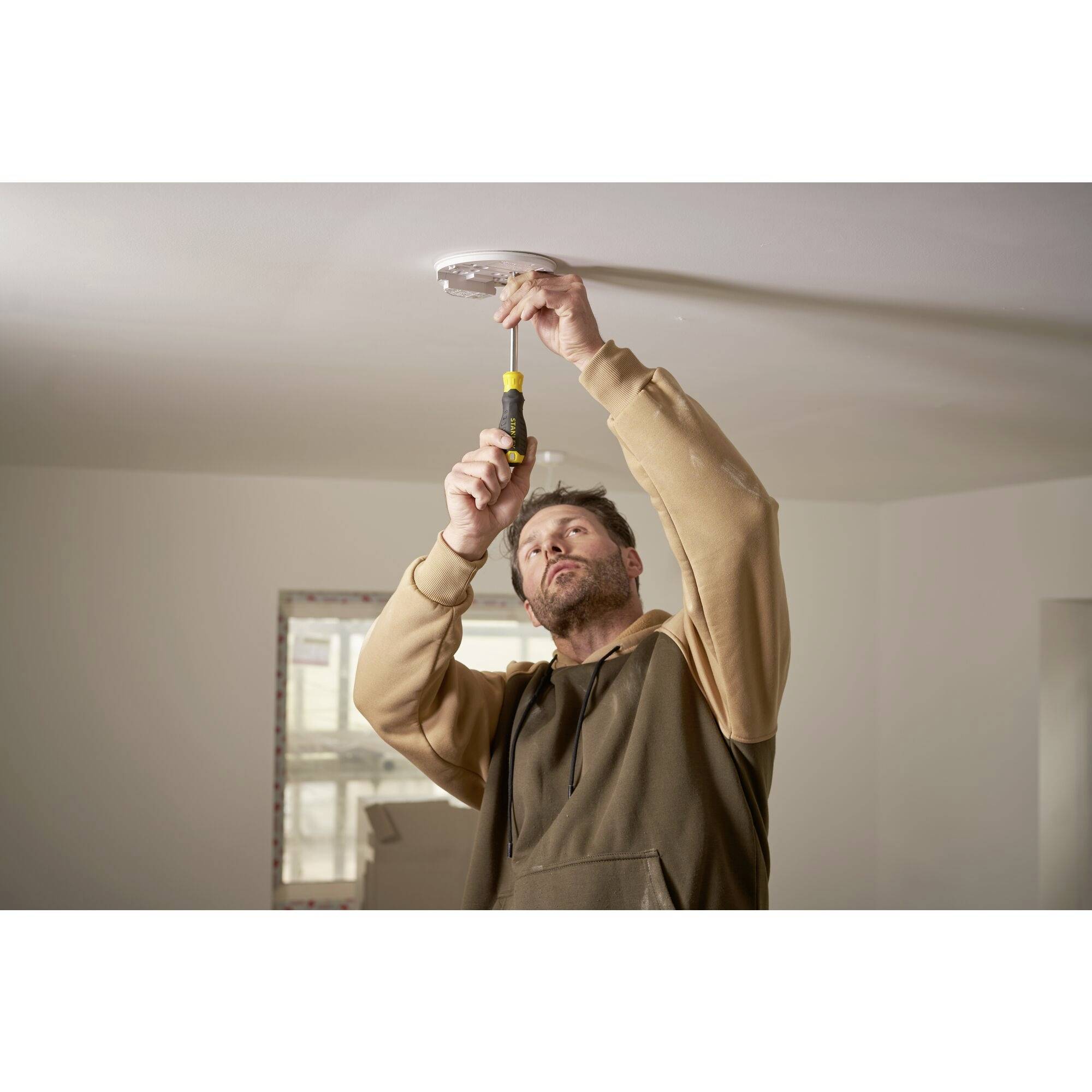 A man is installing a device on a ceiling using a screwdriver. A window can be seen in the background.