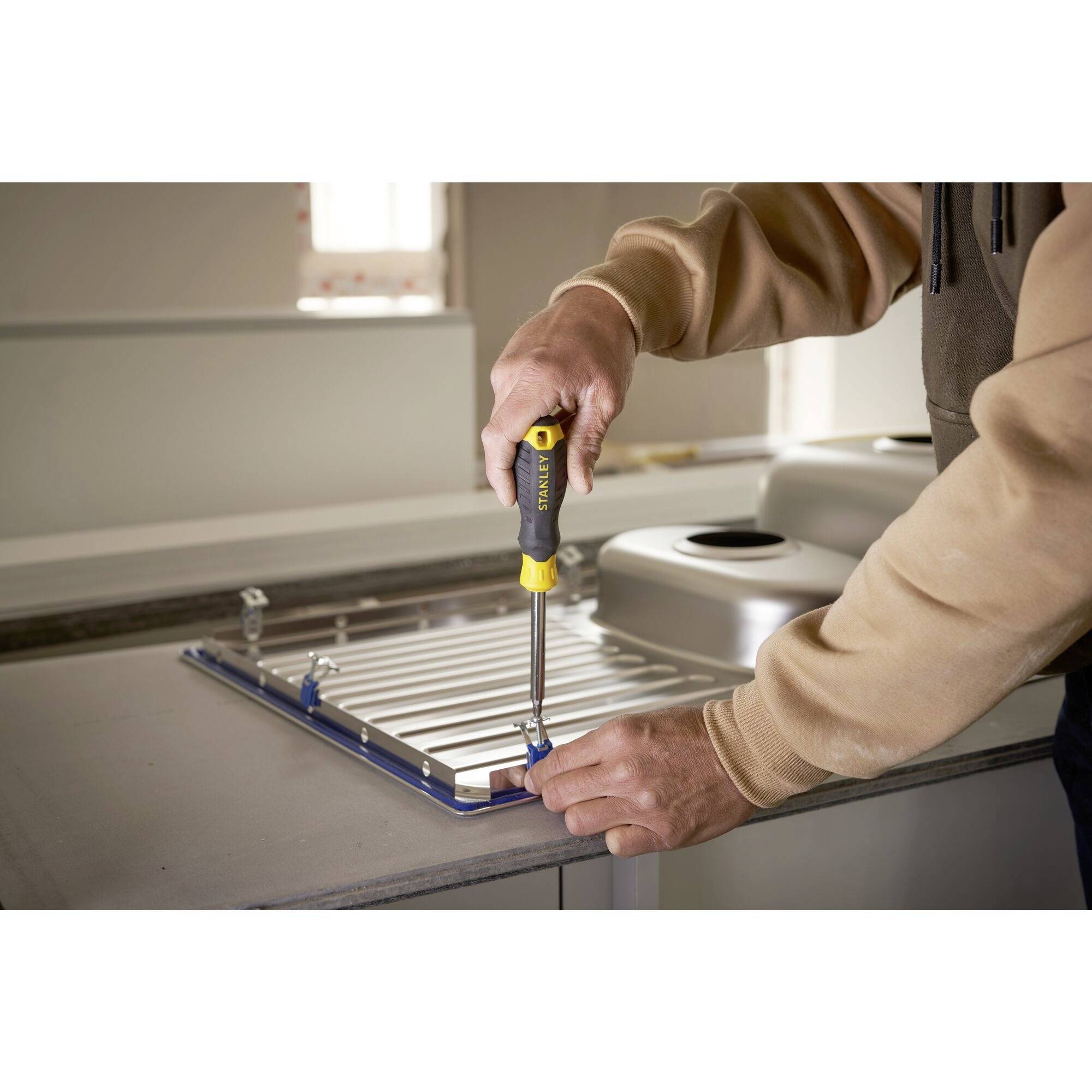 A person is installing a sink on a kitchen worktop with a screwdriver. Kitchen cupboards are visible in the background.
