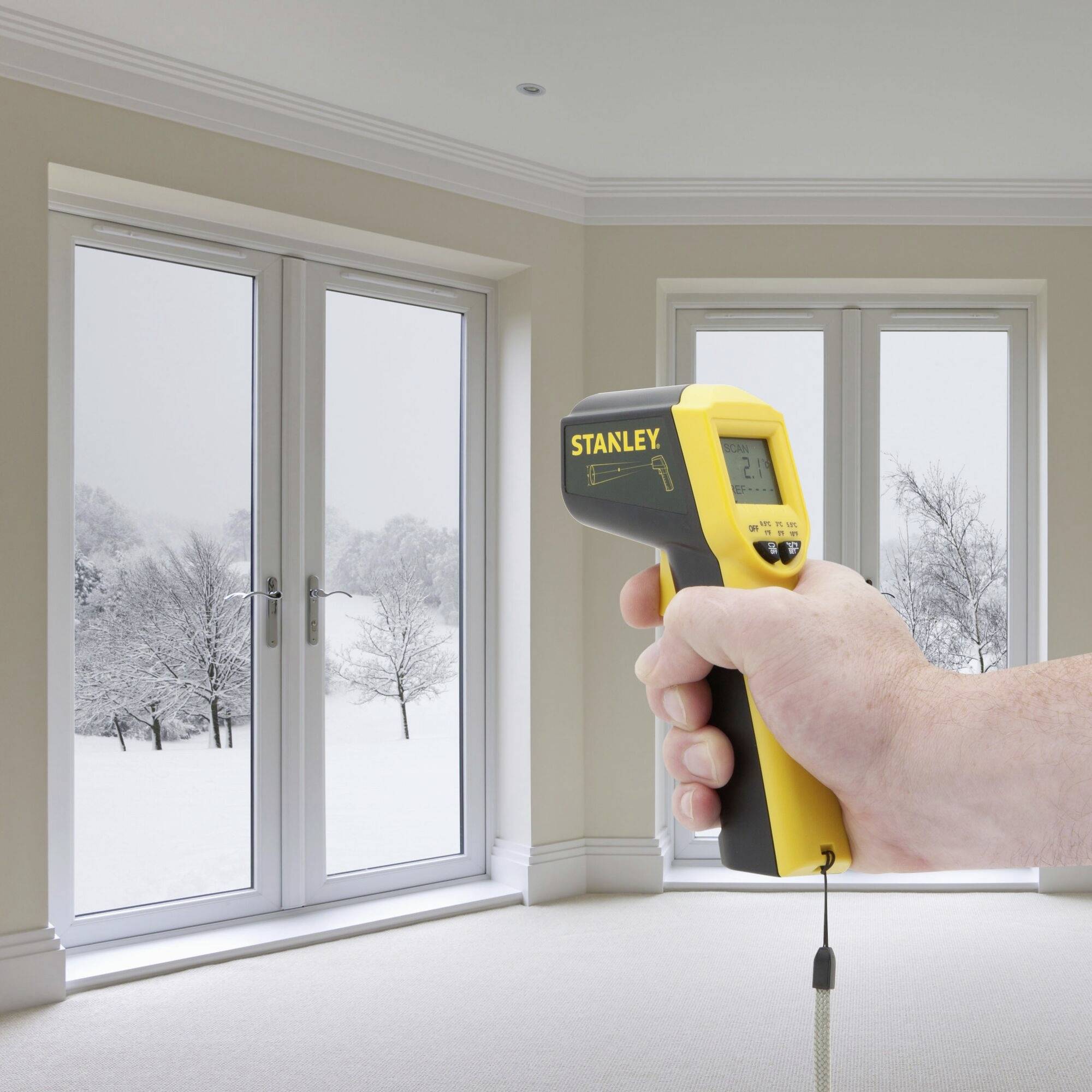 A hand is holding an infrared thermometer in a room with a snow-covered landscape visible through the window. A temperature measurement is being taken.