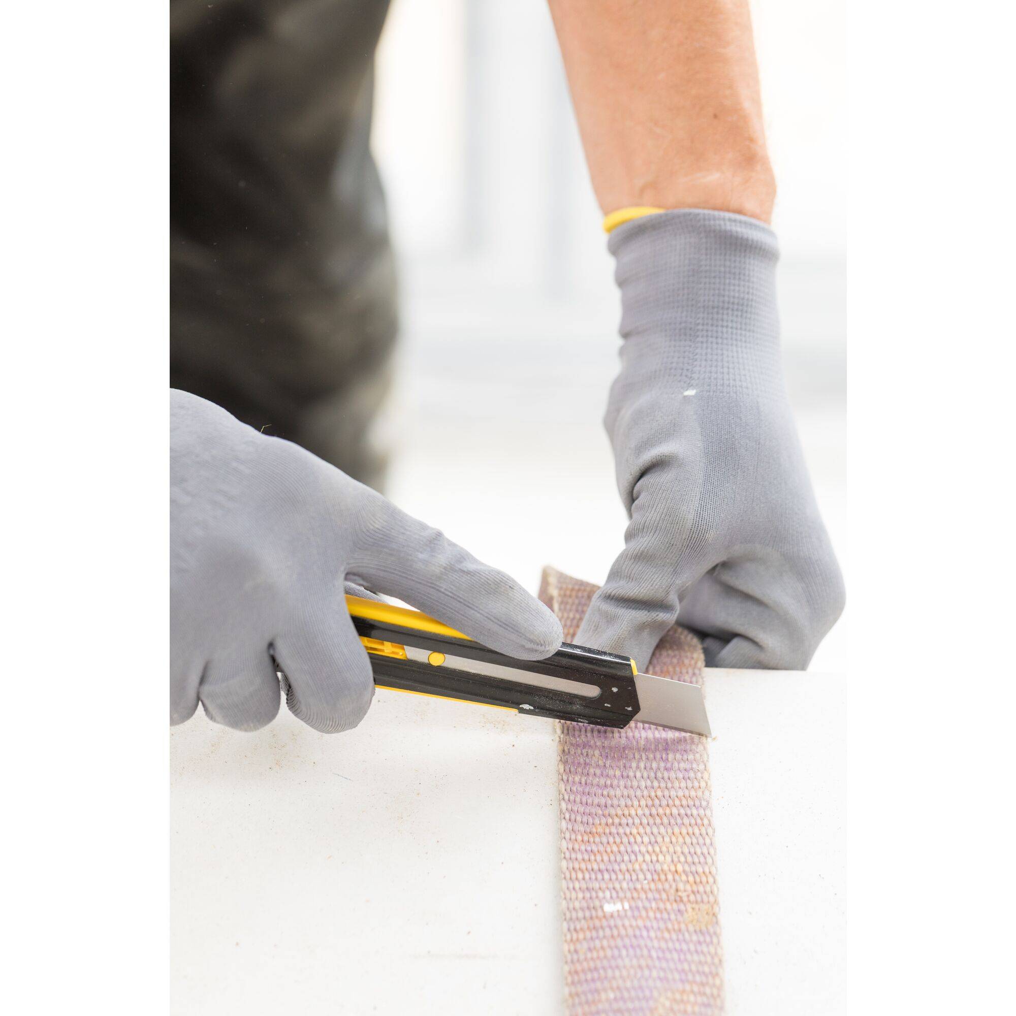 A person wearing grey gloves is cutting a piece of sandpaper with a craft knife.