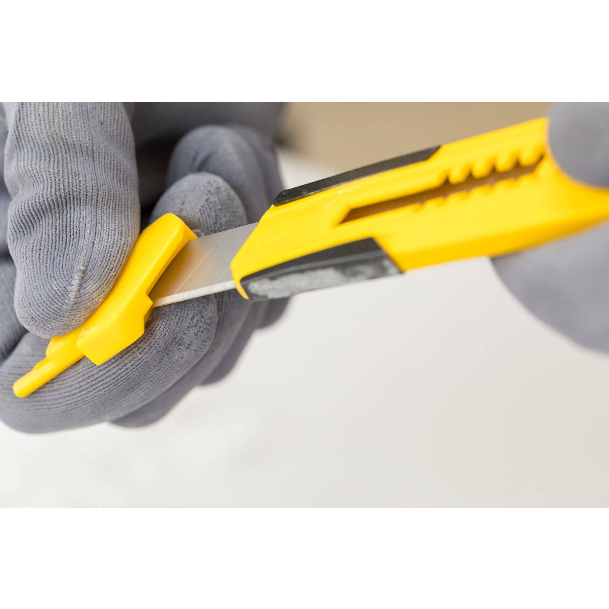 Two hands wearing grey gloves are holding a yellow craft knife that is partially open.
