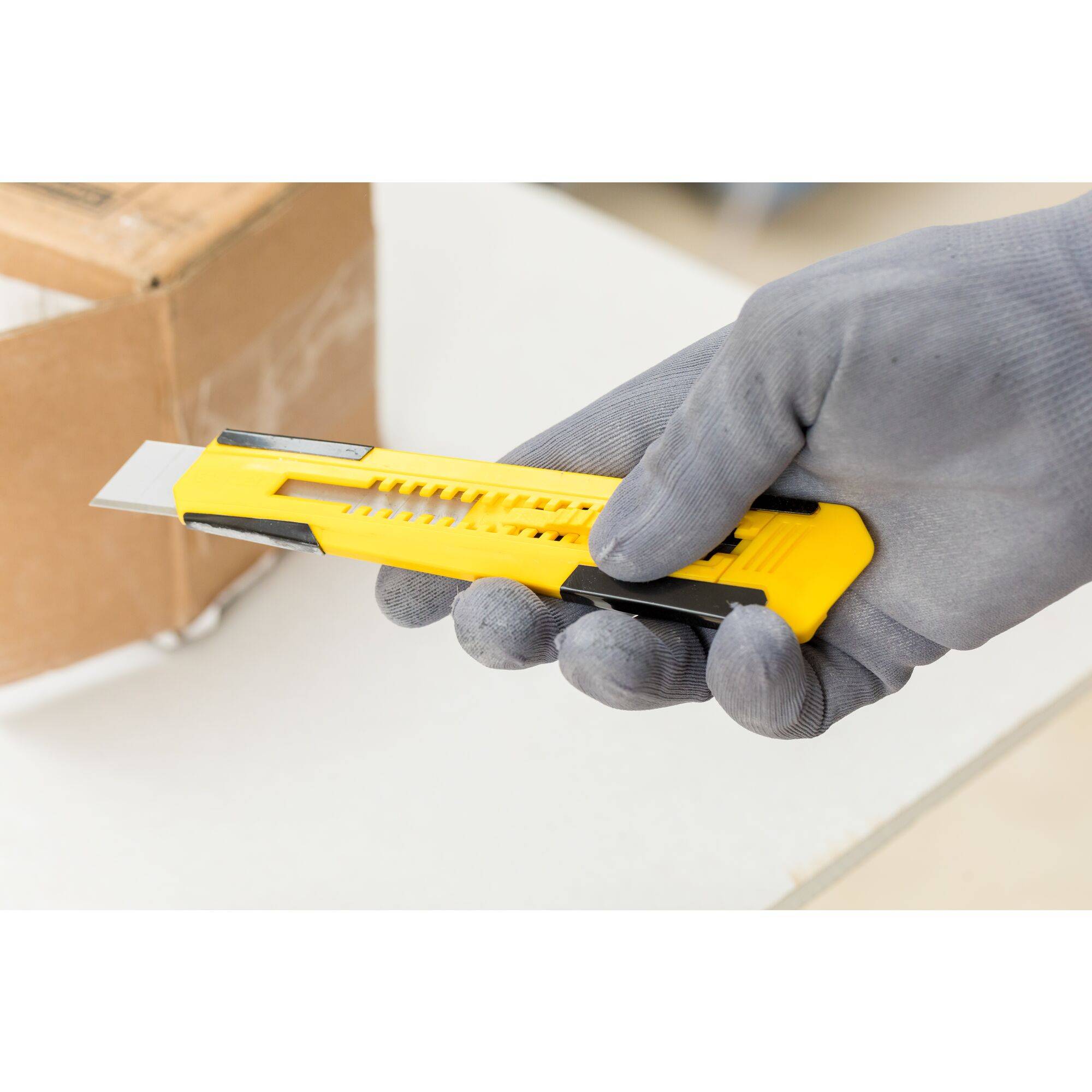 A gloved hand holds a yellow carpet knife, ready to open a cardboard package.
