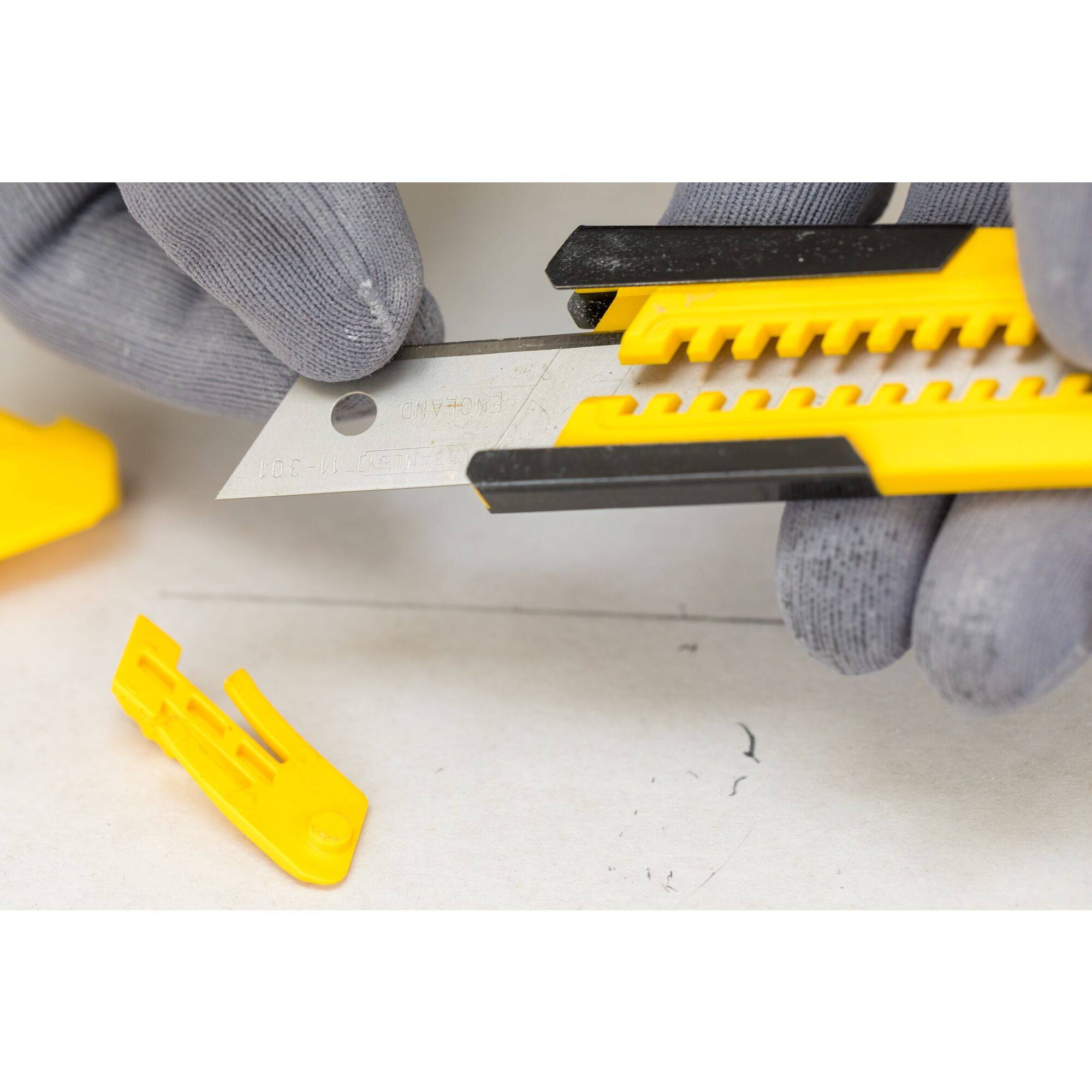 Two hands wearing work gloves are holding a yellow Stanley knife with the blade exposed. A yellow cover is lying beside it.