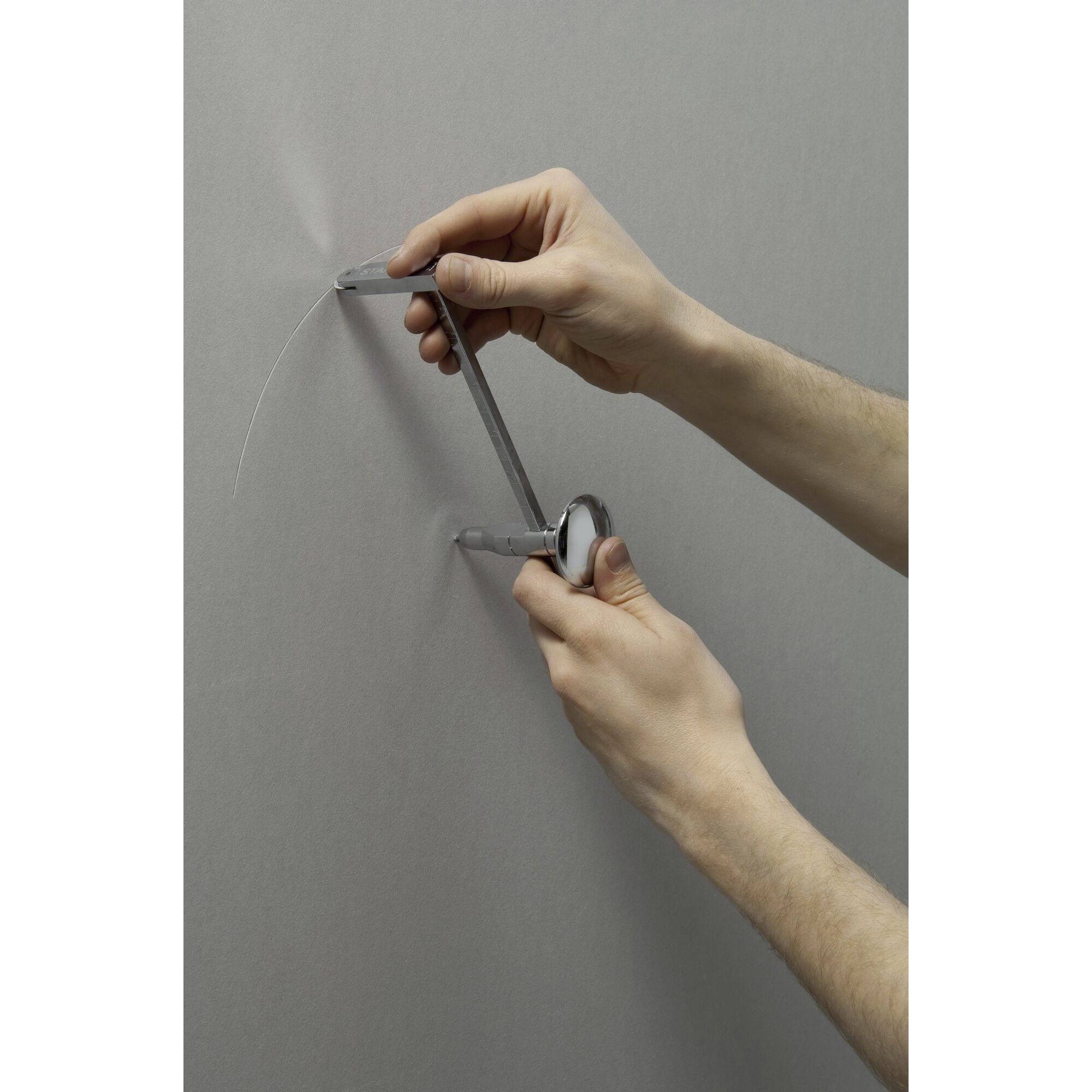 'Two hands hold a compass against a grey wall, drawing a circular line, presumably to mark out a guide.'