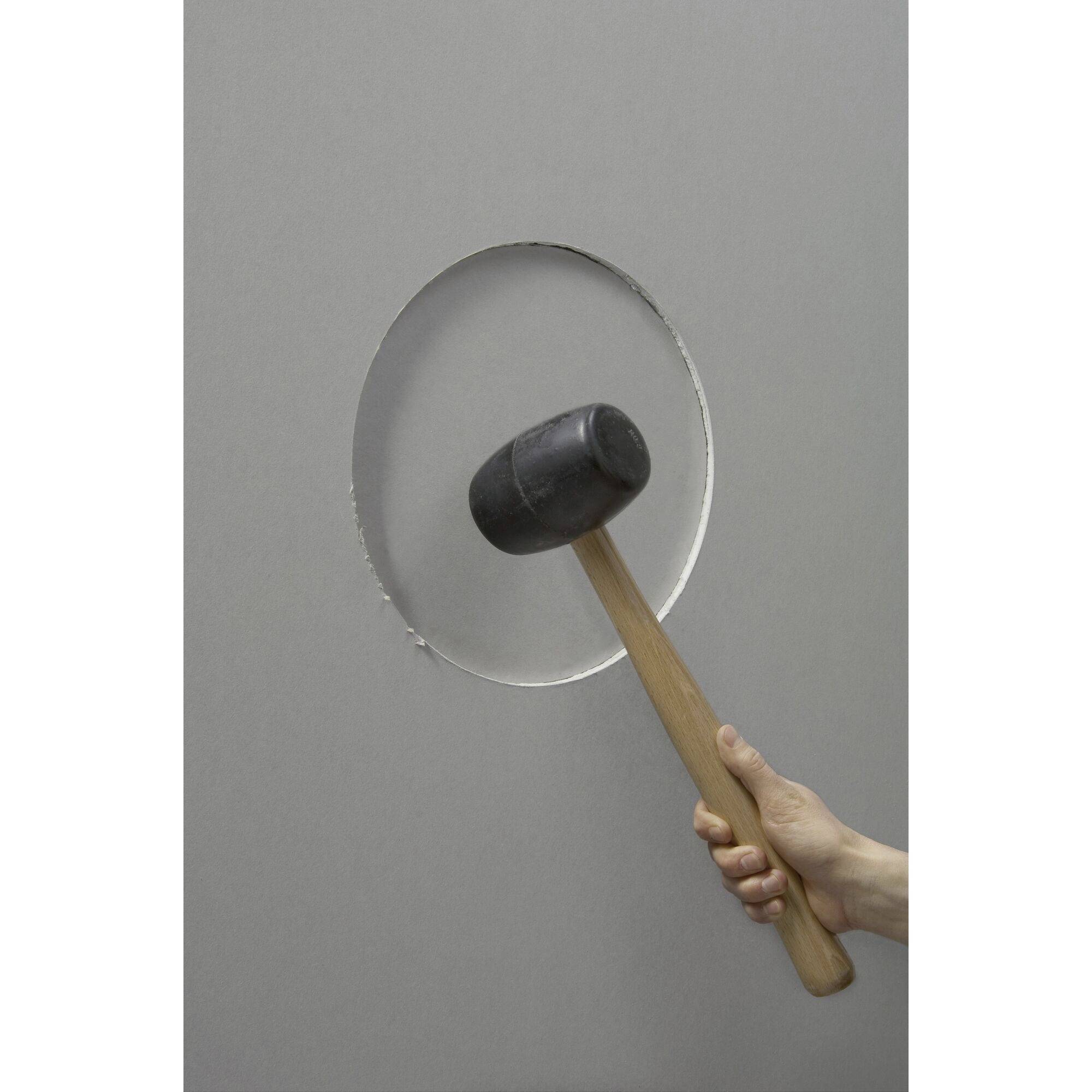 A hand holds a hammer striking against a grey wall, creating a circular hole.