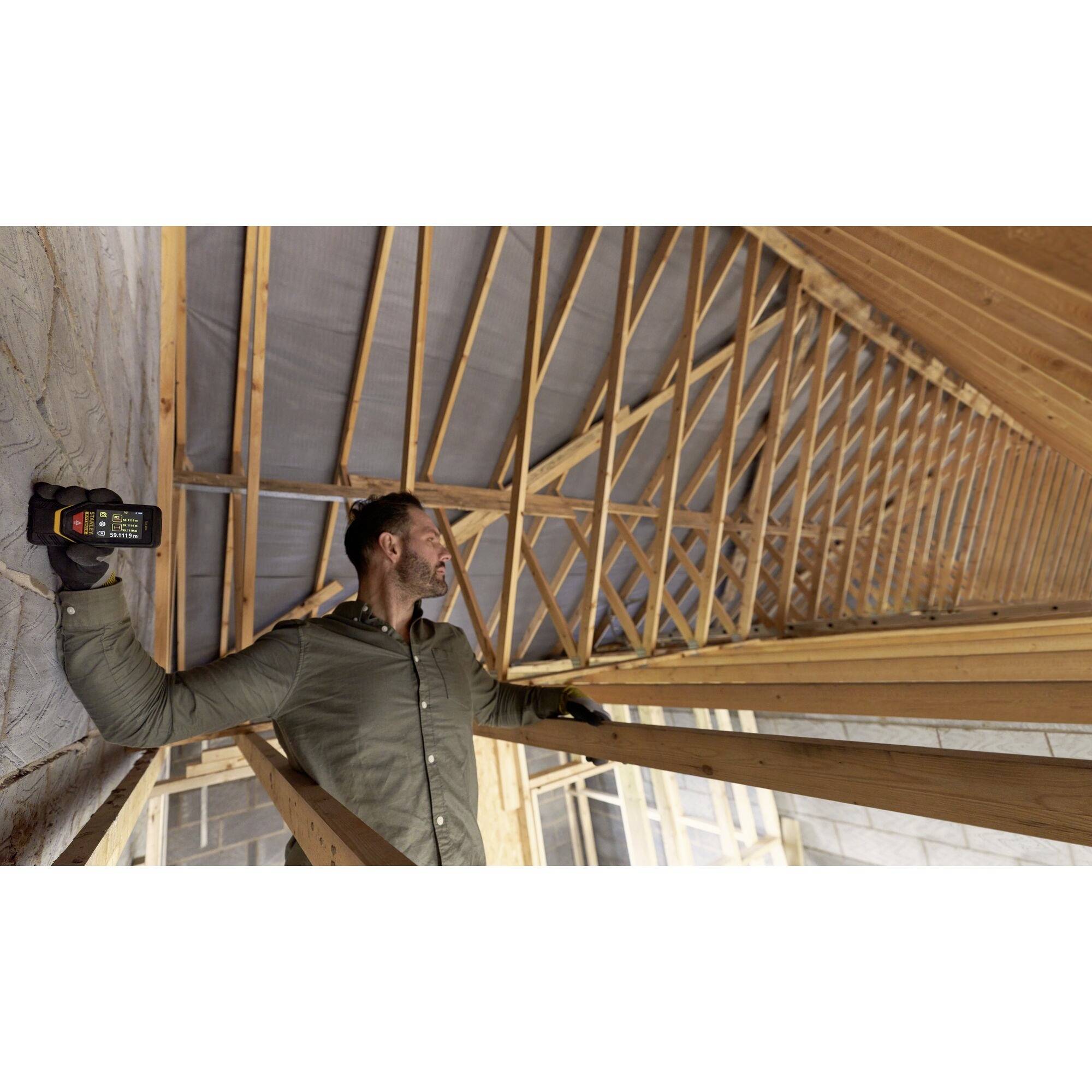 A man on a construction site is checking the interior of a wooden roof structure with a measuring device. He is wearing warm clothing and gloves.