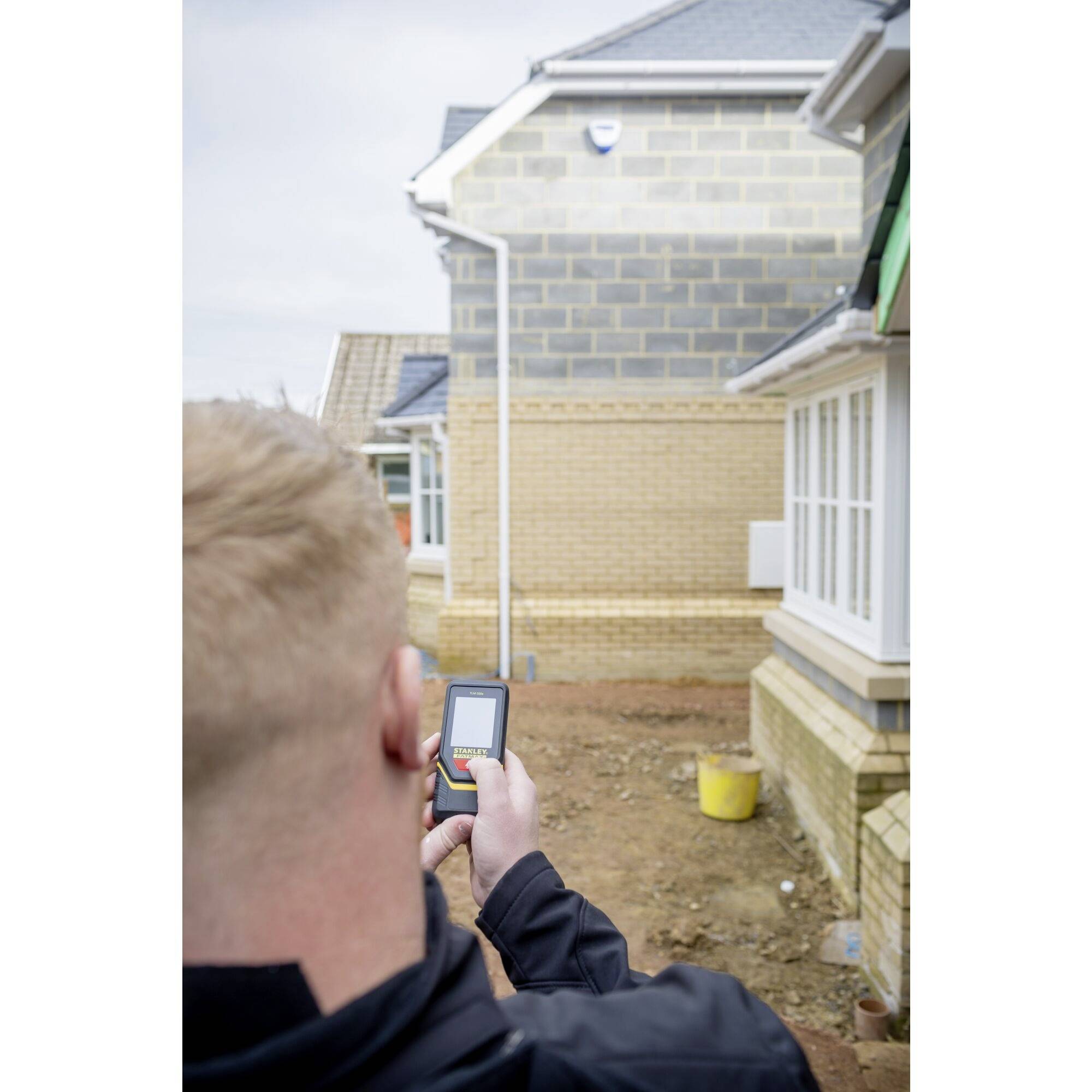 A person is holding a laser distance measuring device and measuring the distance to a partially completed brick house on a construction site.