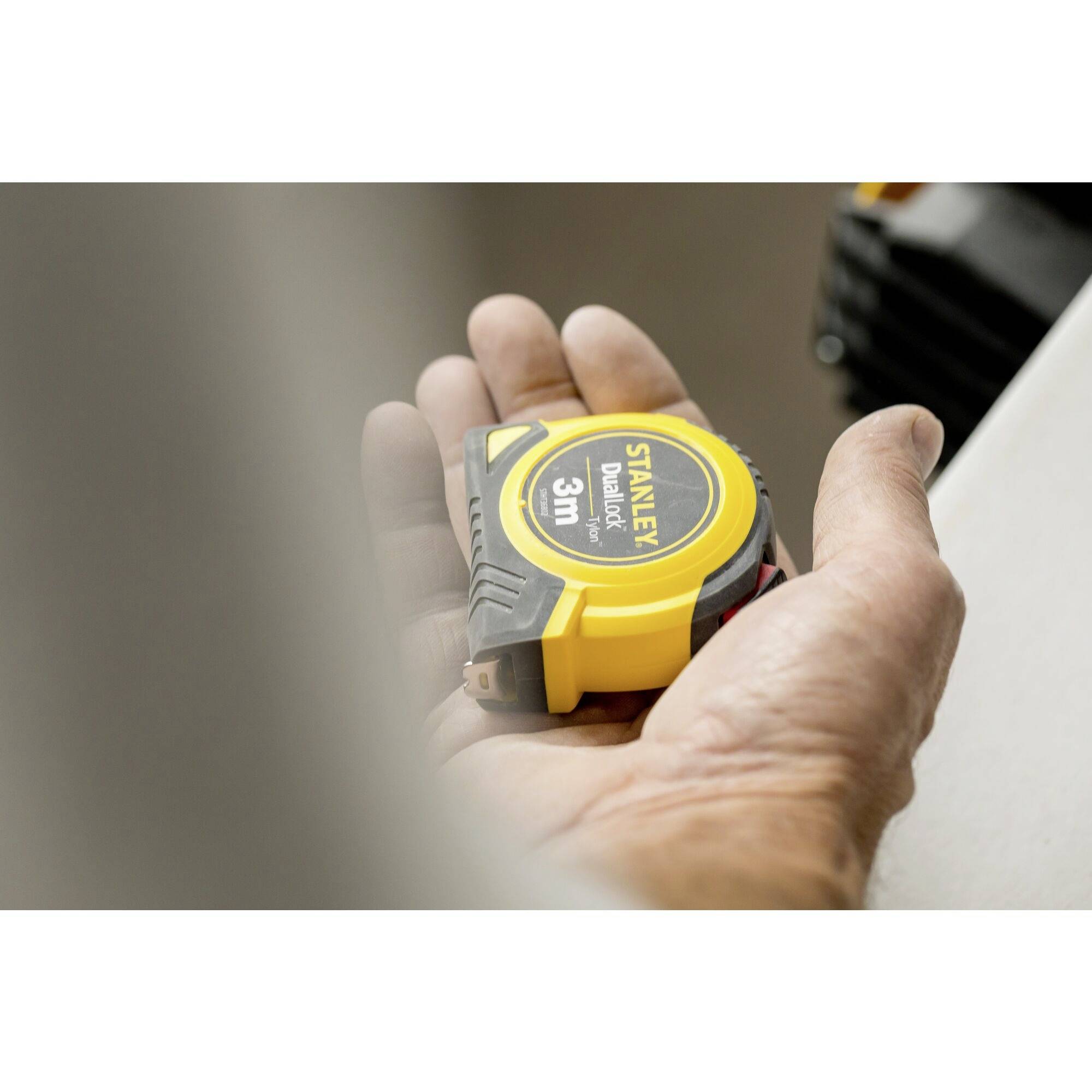 A hand is holding a yellow tape measure. On the front of the tape measure, it reads 'Stanley Dual Lock, 3m'.