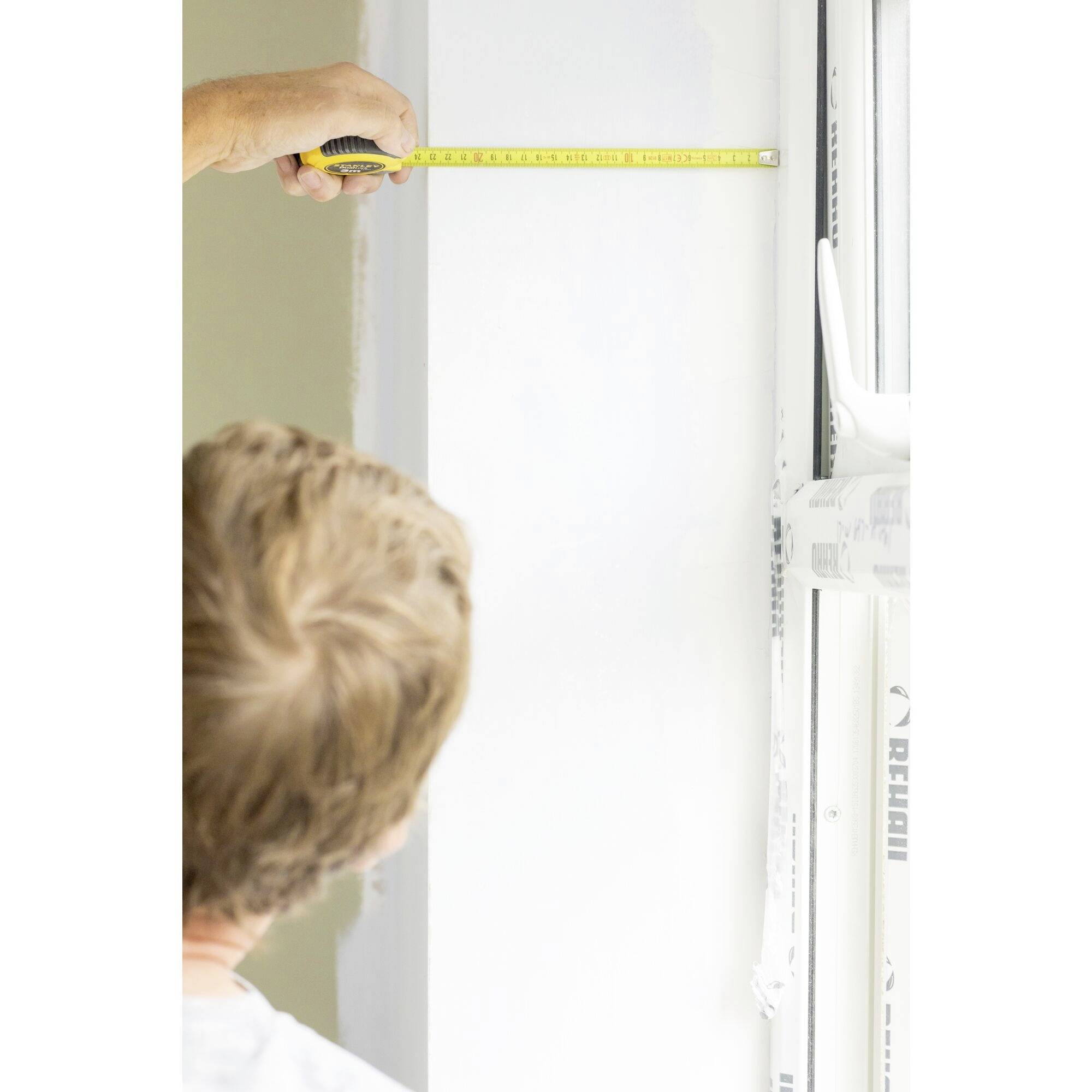 A person is measuring the width of a white wall with a tape measure, focusing on precision during construction work.