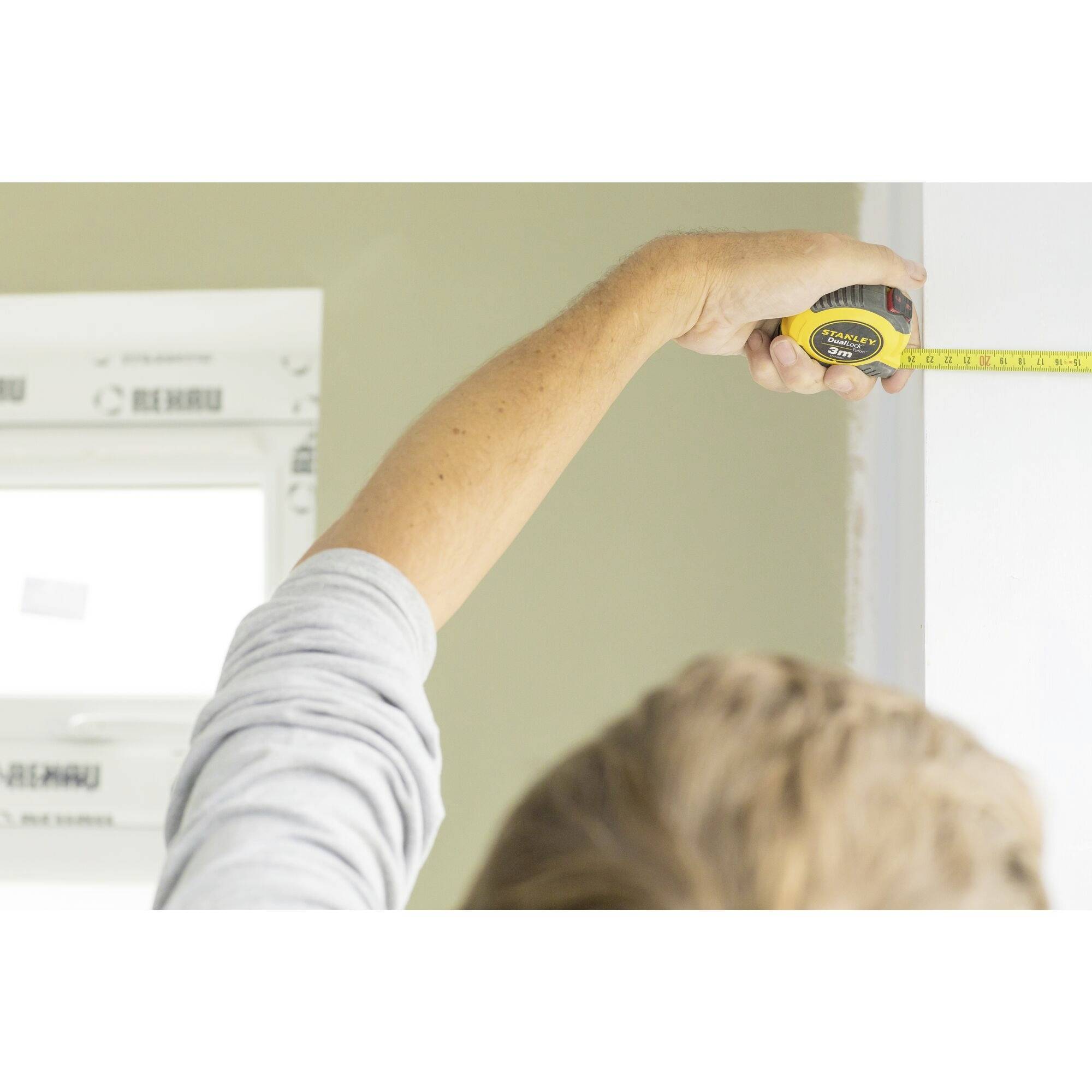 A person is measuring the width of a wall with a measuring tape. Part of a window frame is visible in the background.