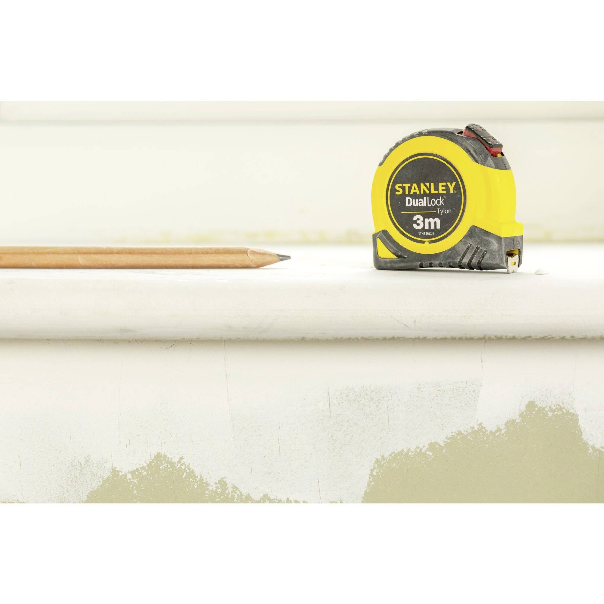 A yellow measuring tape with the inscription 'Stanley DualLock 3m' lies beside a pencil on a white table.