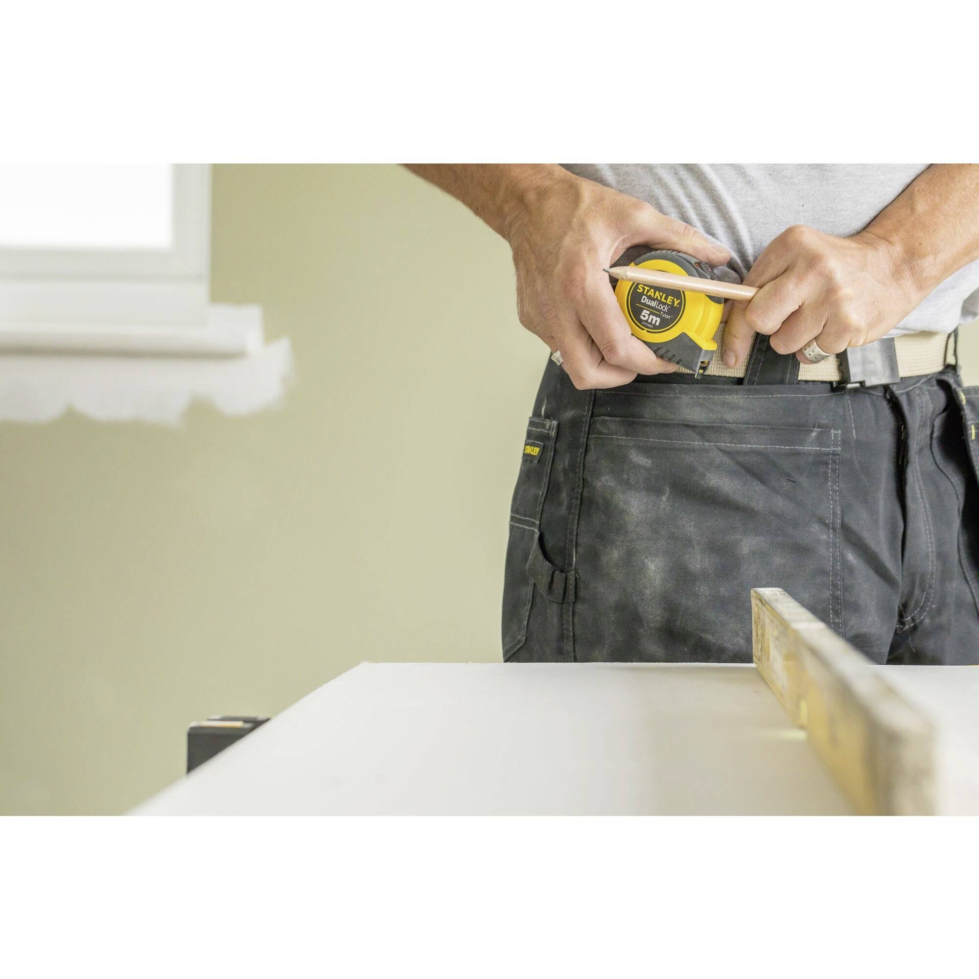 A person is putting a tape measure into their trouser pocket. A table is visible in the foreground. The focus is on practical work.