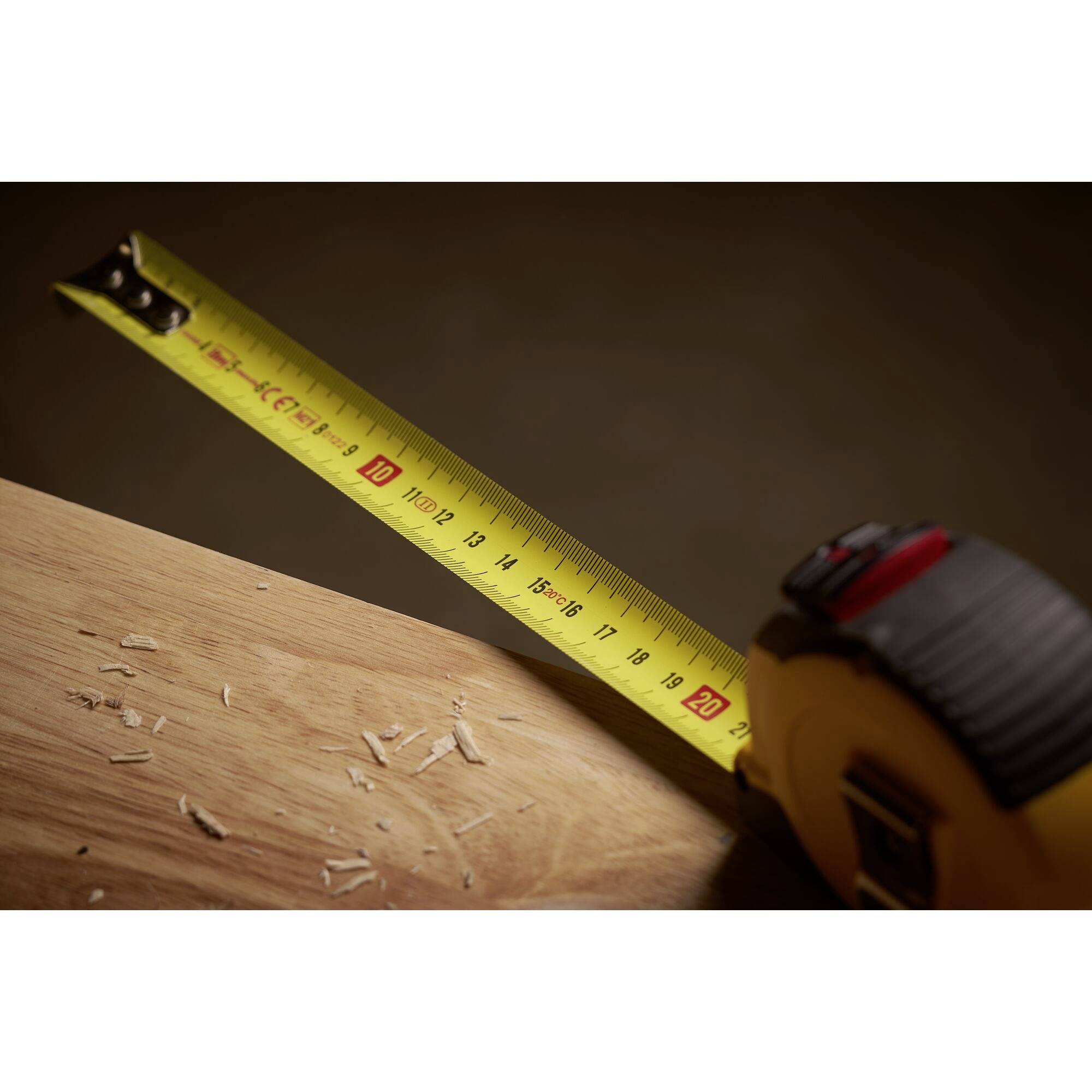 A yellow tape measure lies on a wooden table, selected length at 20 cm. Wood shavings are scattered across the table surface.