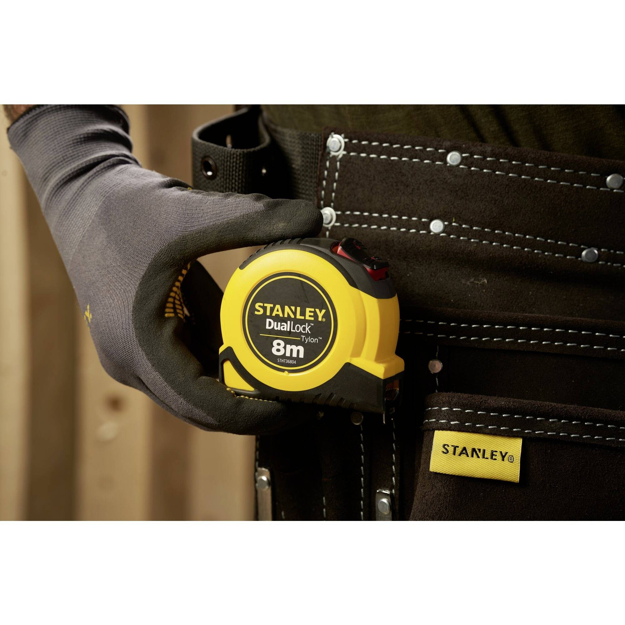 A person wearing work gloves is holding a Stanley tape measure, 'DualLock 8m', on a dark tool belt.