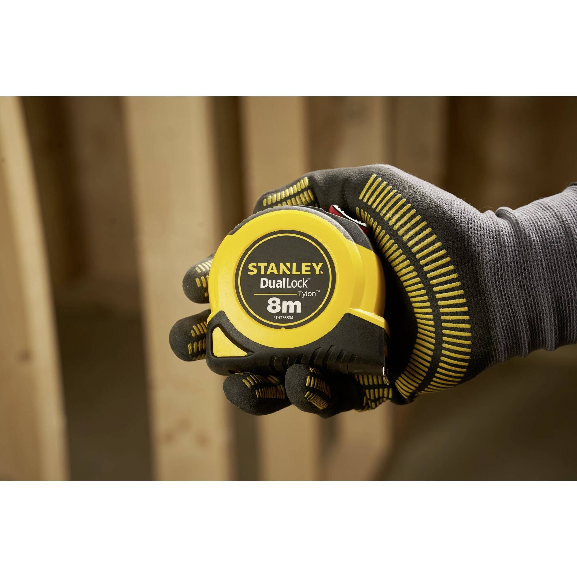 A gloved hand holds a yellow Stanley DualLock measuring tape with 8 metres in length. The background shows blurry wooden boards.