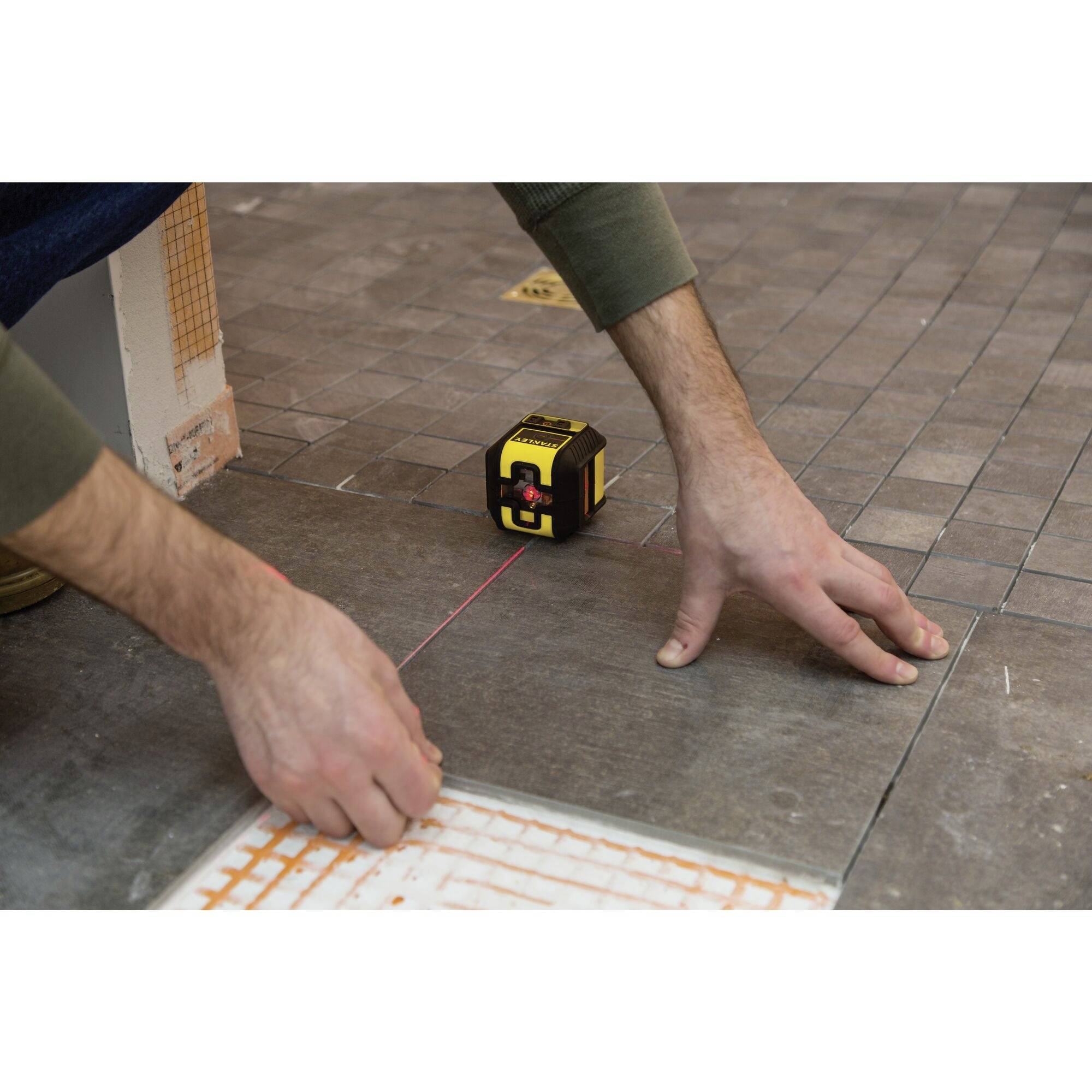 A person is aligning tiles with a laser measuring device on a floor. The device is projecting a red laser line for alignment.