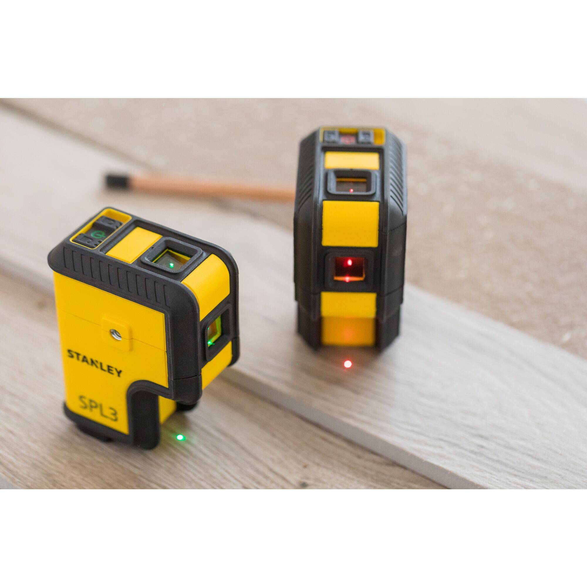 Two yellow and black laser levelling devices on a wooden floor. One is showing red laser beams, the other green. A pencil in the background.