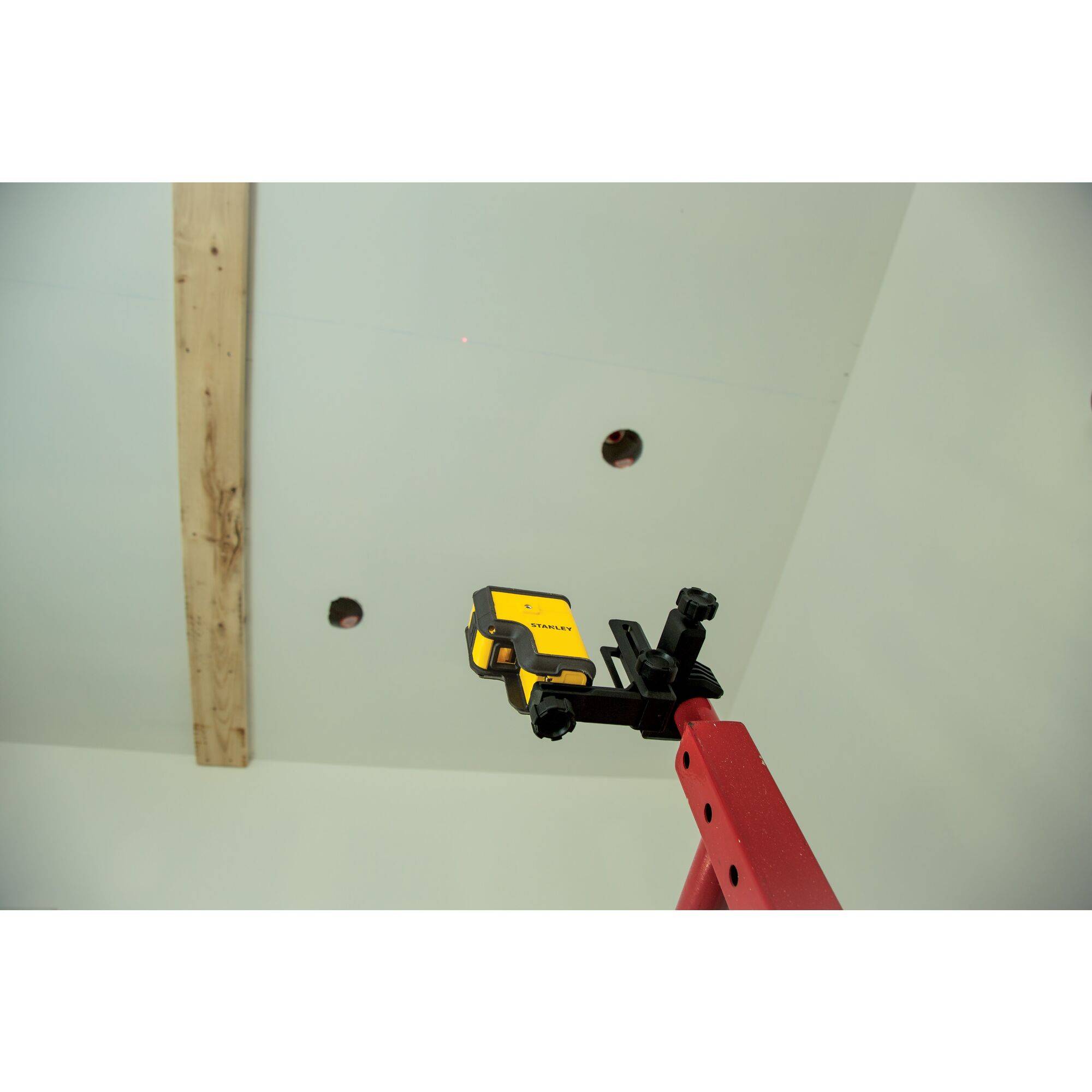 A yellow laser level mounted on a red tripod is aimed at two drill holes in a ceiling, next to a wooden beam.