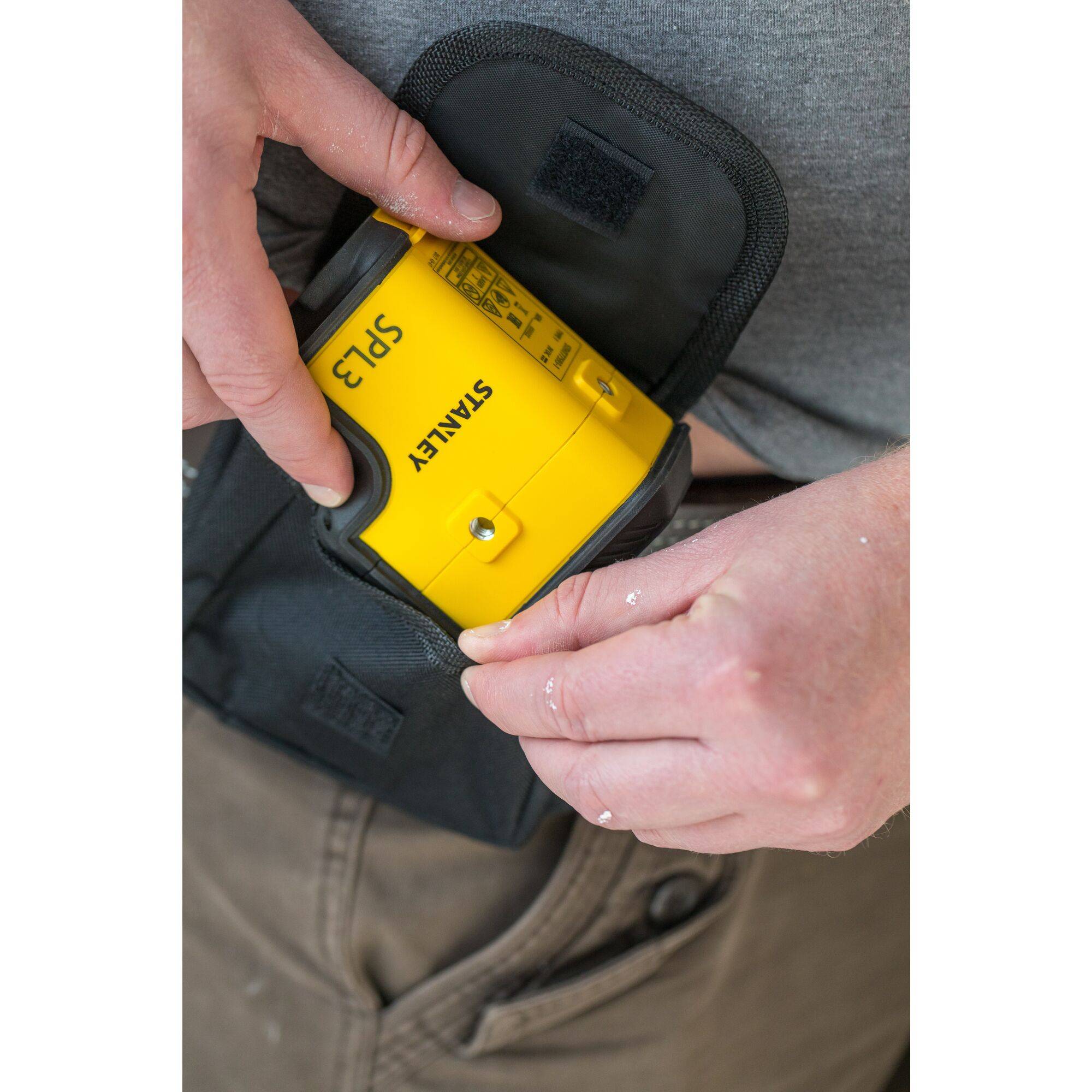 A person is putting a yellow tool with the inscription 'STANLEY SP2' into a black bag. Hands have white paint splatters.