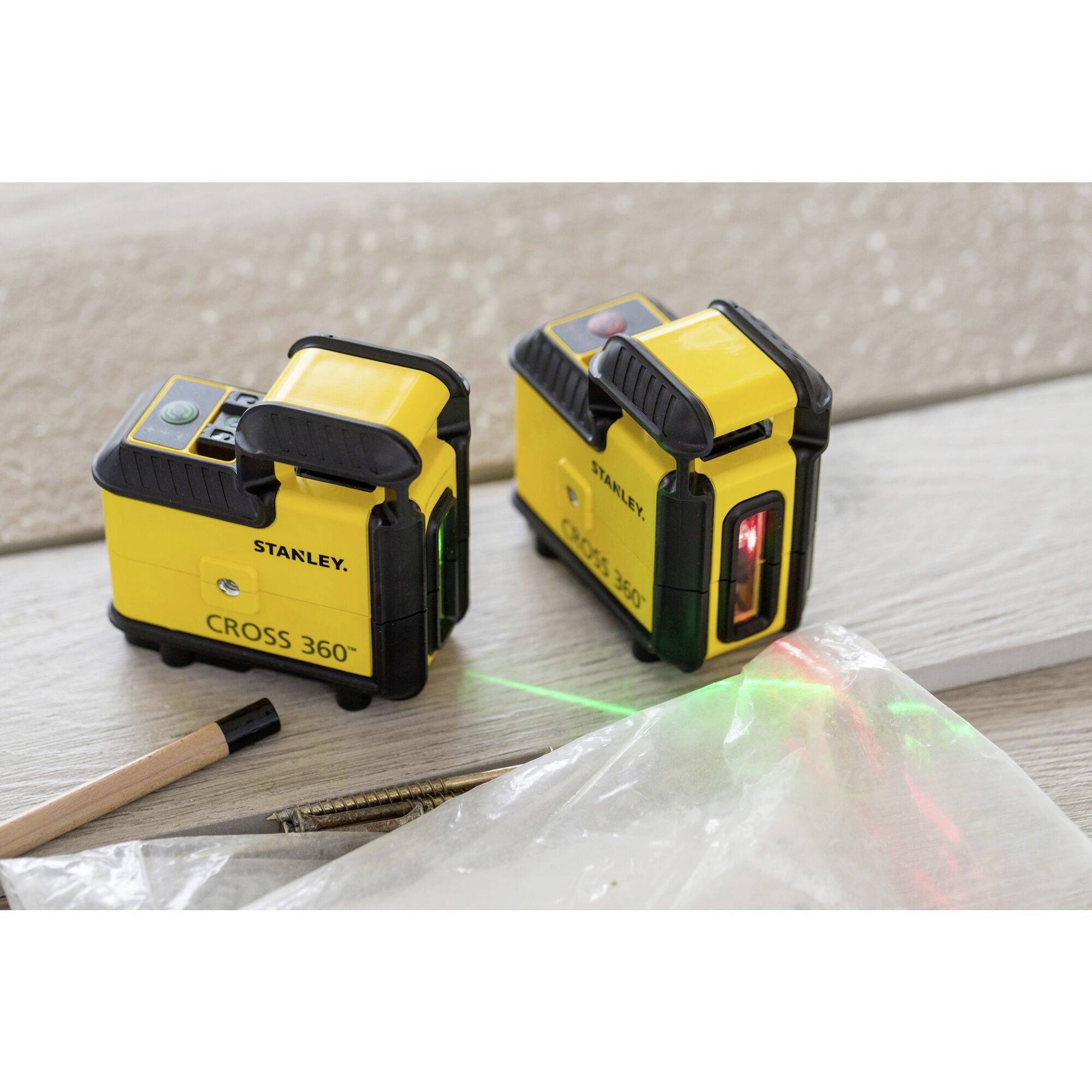 Two yellow laser spirit levels on a wooden floor; one projecting red and green lines. In the foreground, a pencil and screws.