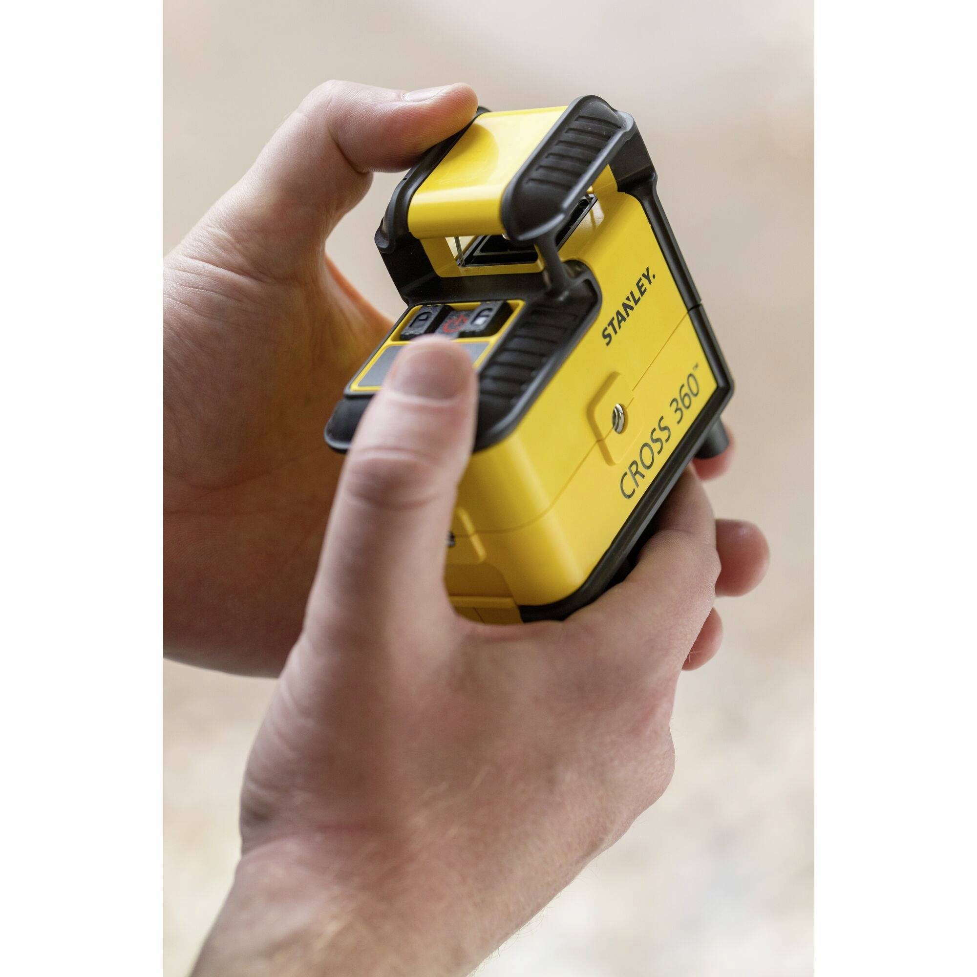 A person is holding a yellow, square device with the print 'CROSS 360' and the brand name. The device appears to be a tool.