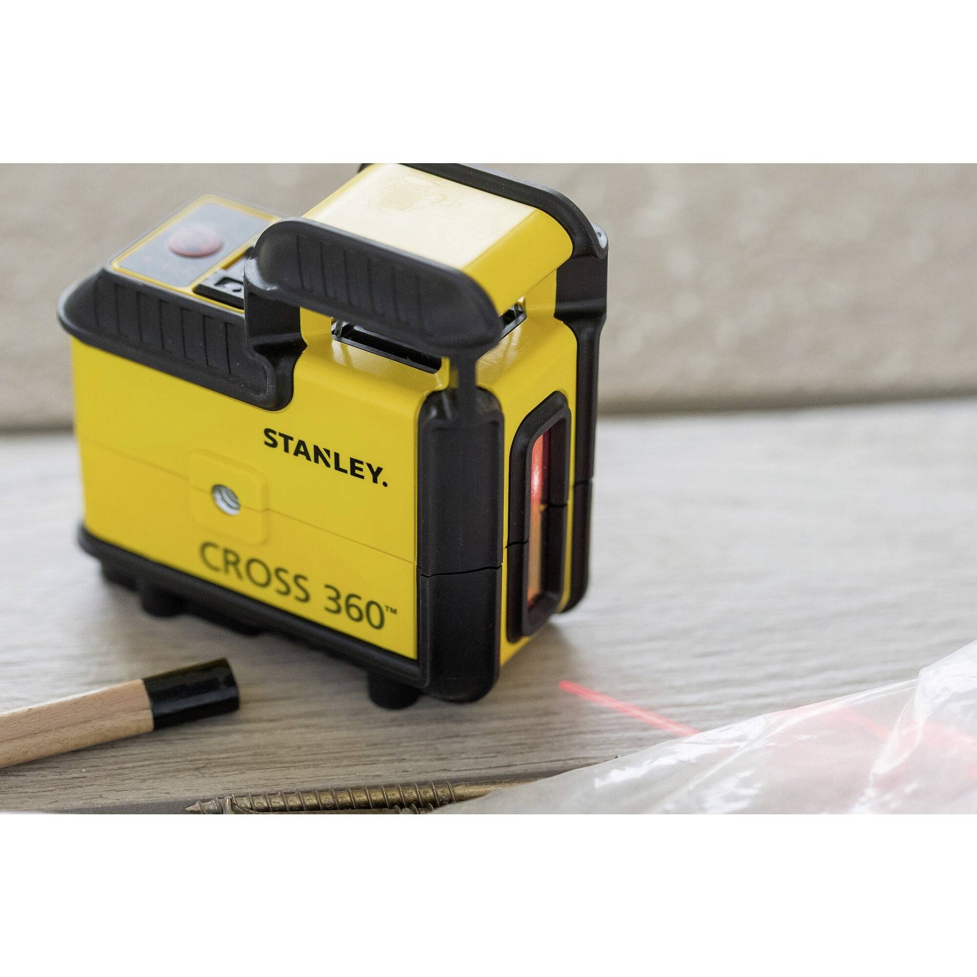 A yellow cross-line laser device with the inscription 'Stanley Cross 360°', positioned on a wooden floor. A pen and screws are lying beside it.