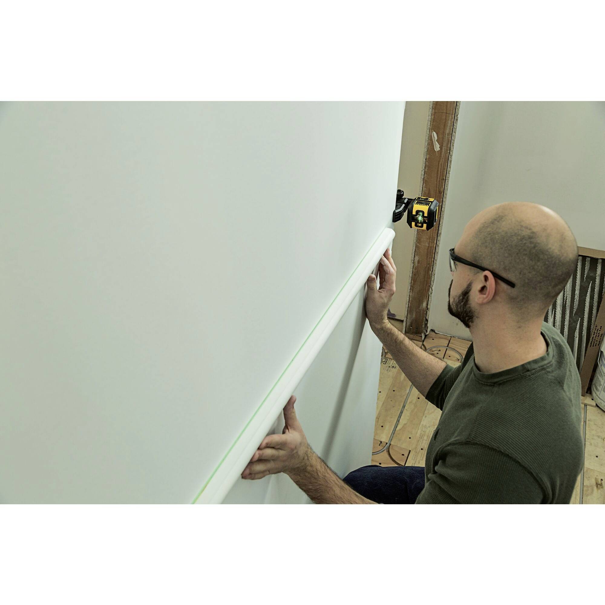 A man is installing a white decorative trim on a wall using a laser measuring device for precise alignment.