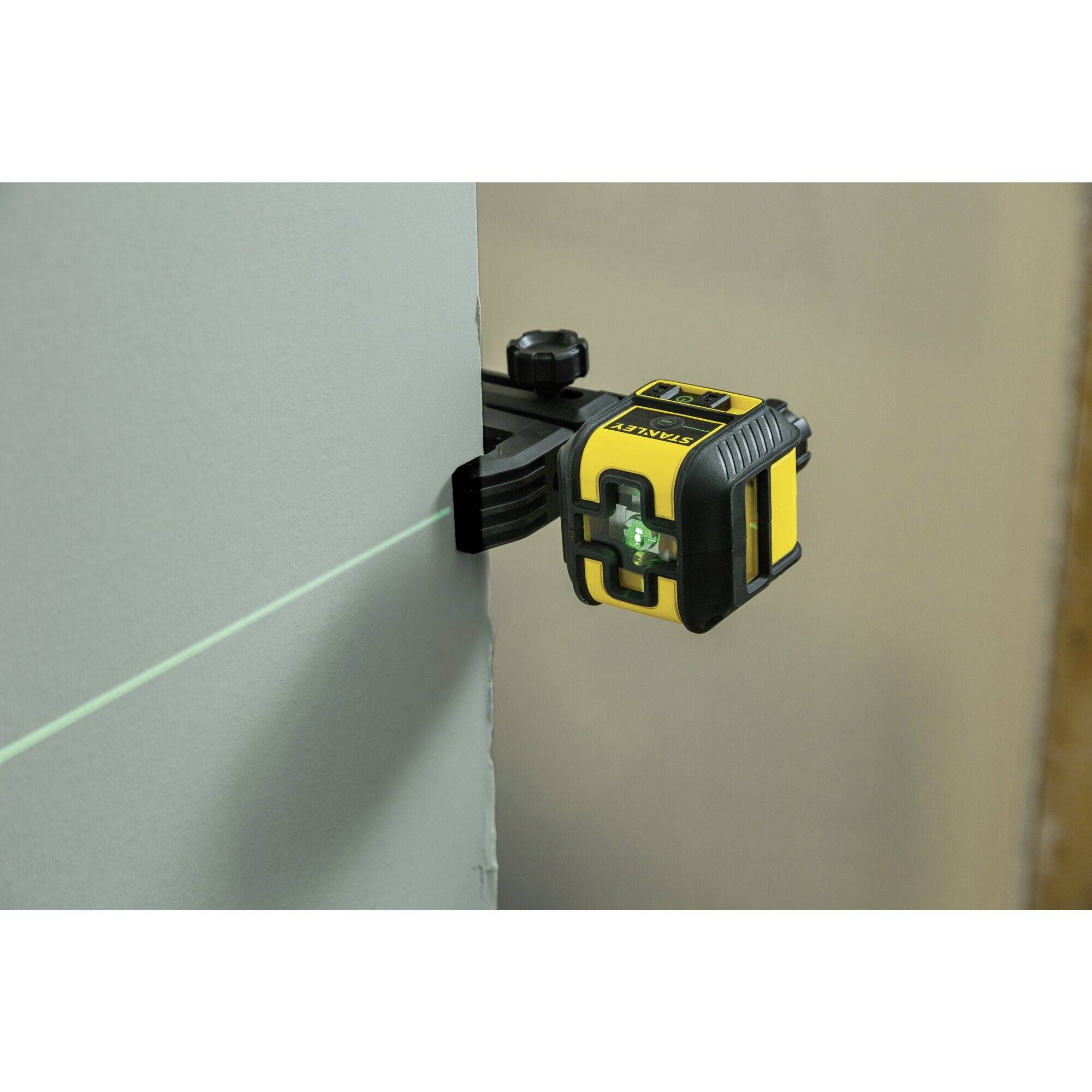 A yellow and black laser levelling device projects a green horizontal line onto a grey wall, showing precise alignment.