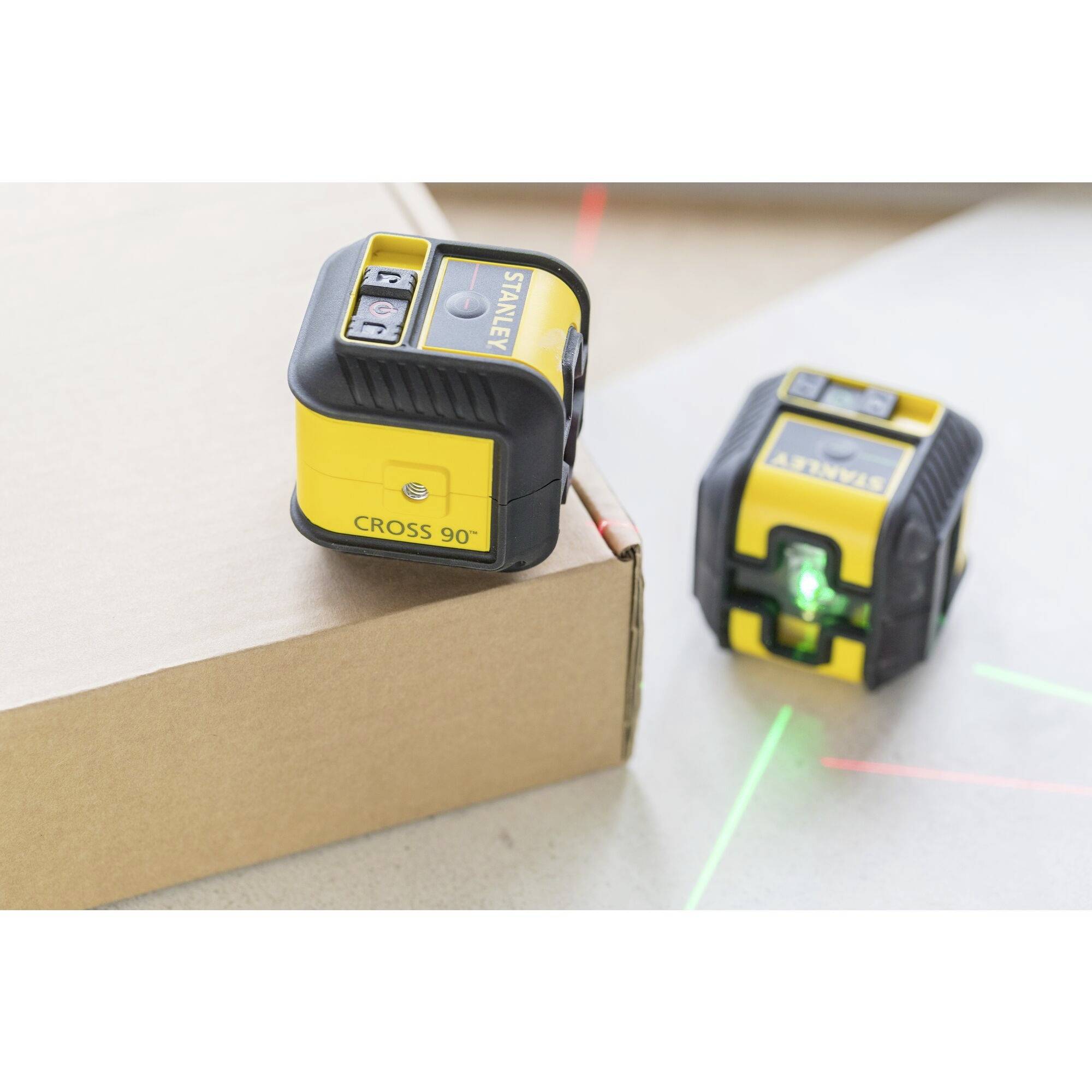 Two yellow-black laser level measuring devices show green and red laser lines on a cardboard box and a flat surface.