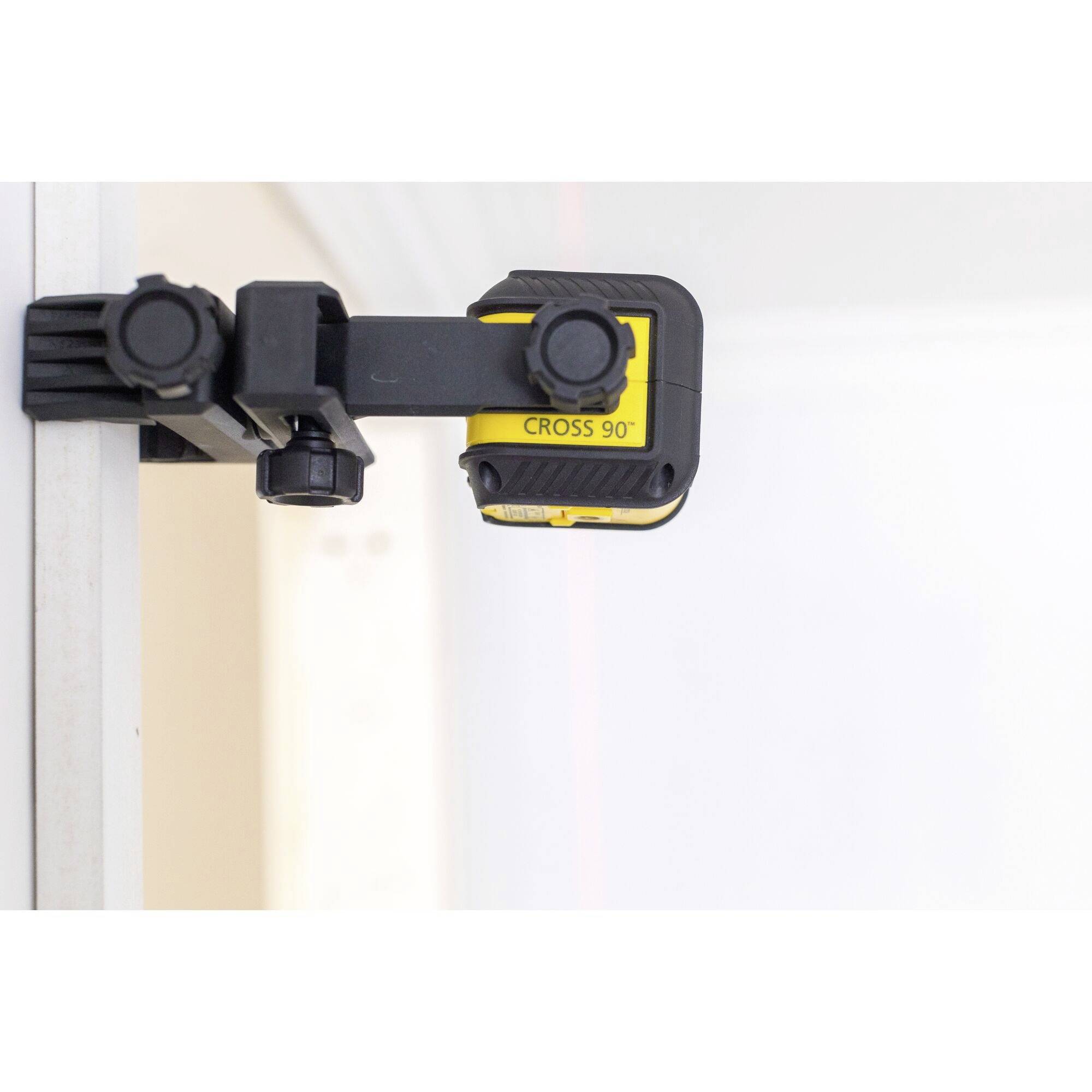 A yellow laser level device with the inscription 'CROSS 90°' is attached to a metal rail and is projecting a red laser beam.