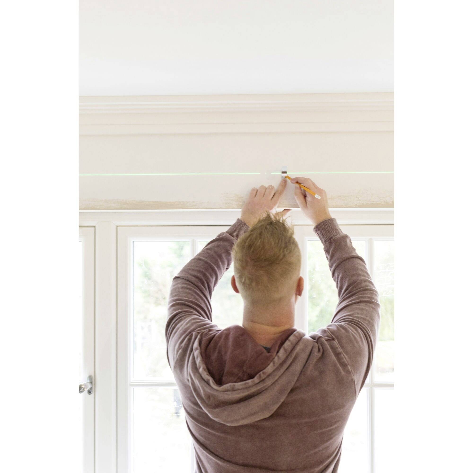 A person is marking a straight line on the wall above a window with a pen. They are wearing a hoodie and standing with their back to the camera.